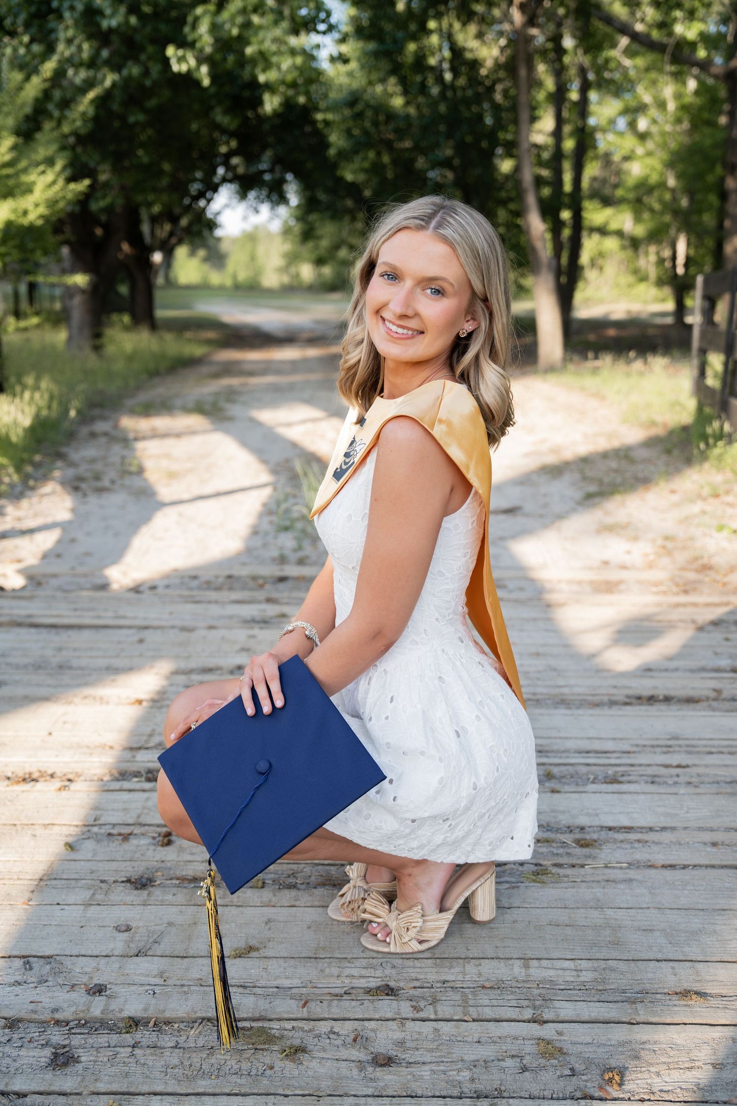 Smiling blonde graduate in white dress holding blue cap on wooden bridge outdoors.