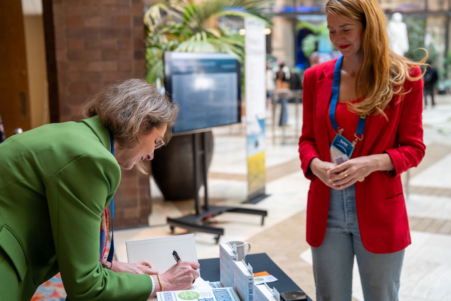 Two people interact at a table in what appears to be a public space, with coffee cups and papers present.