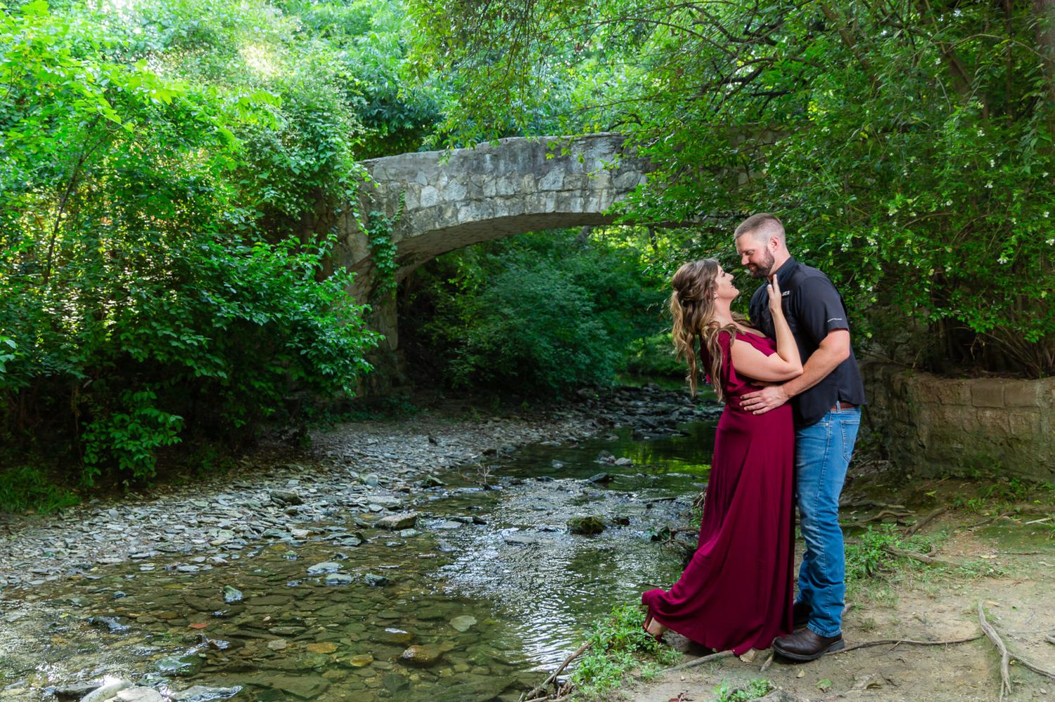 A romantic outdoor engagement photo session along a scenic creek with stone bridge and lush greenery in the background.