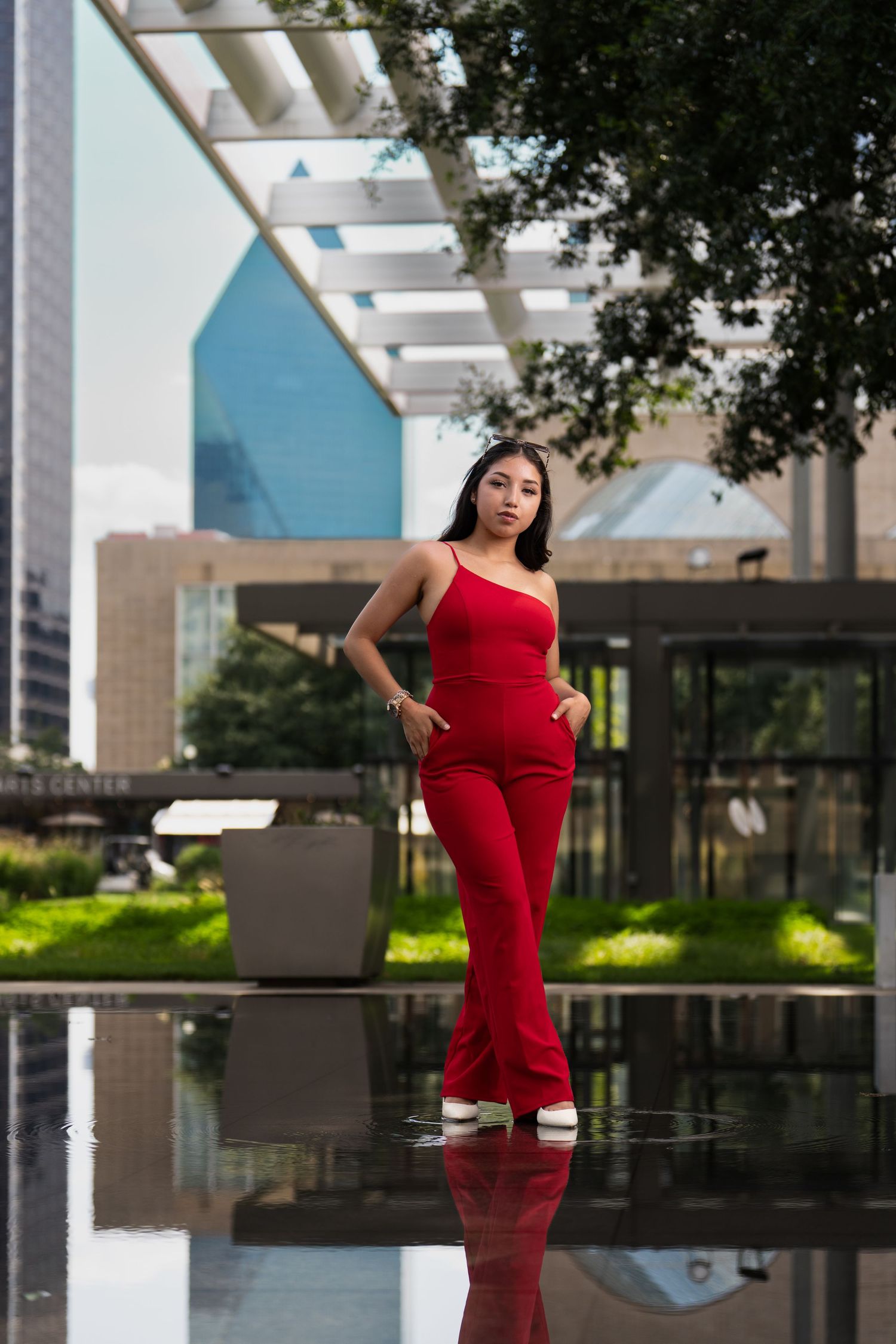 A one-shoulder red jumpsuit outfit photographed in an urban setting with reflective surfaces and modern architecture.