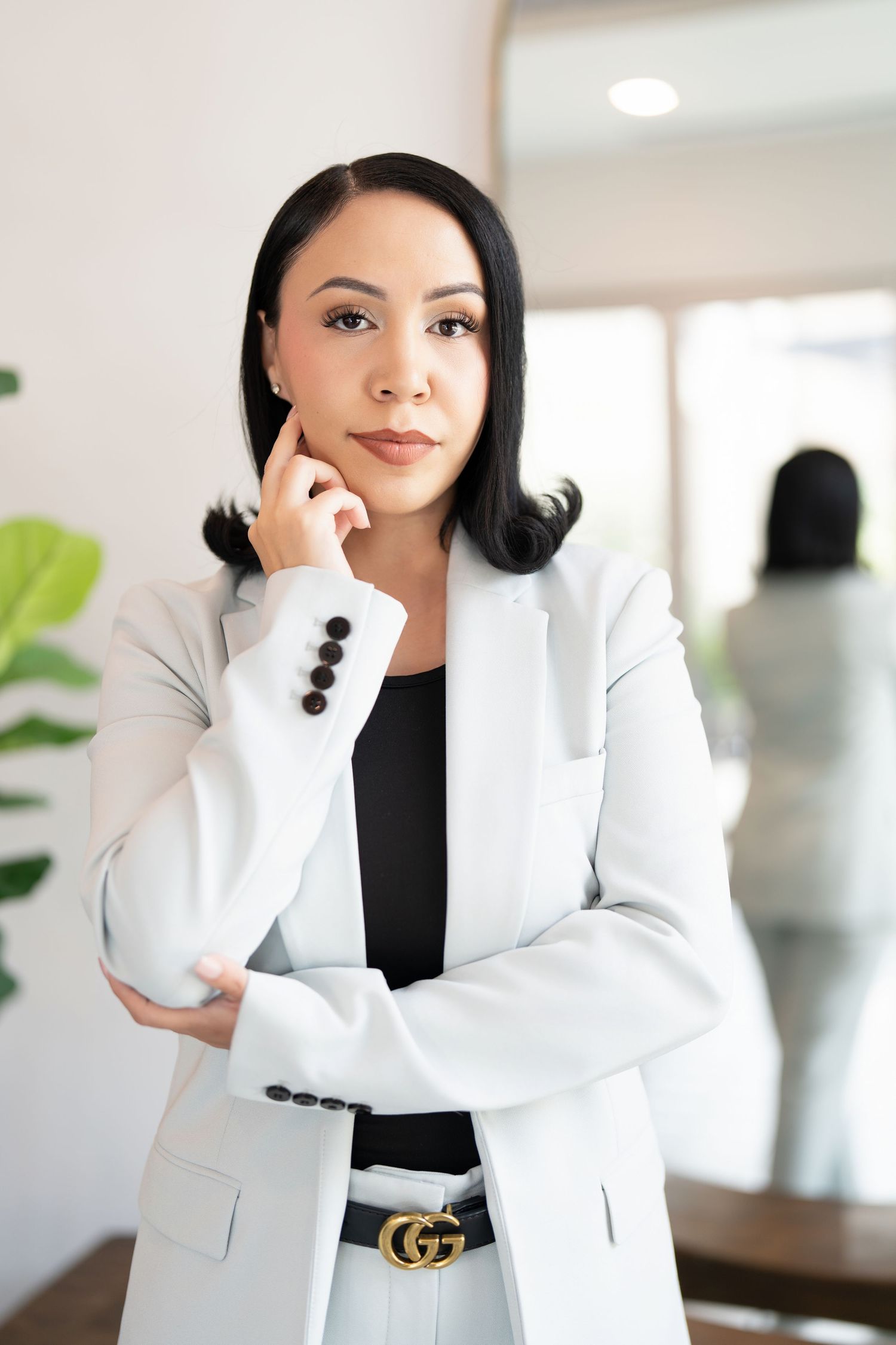 Professional businesswoman in white blazer and designer belt poses confidently in modern office setting.