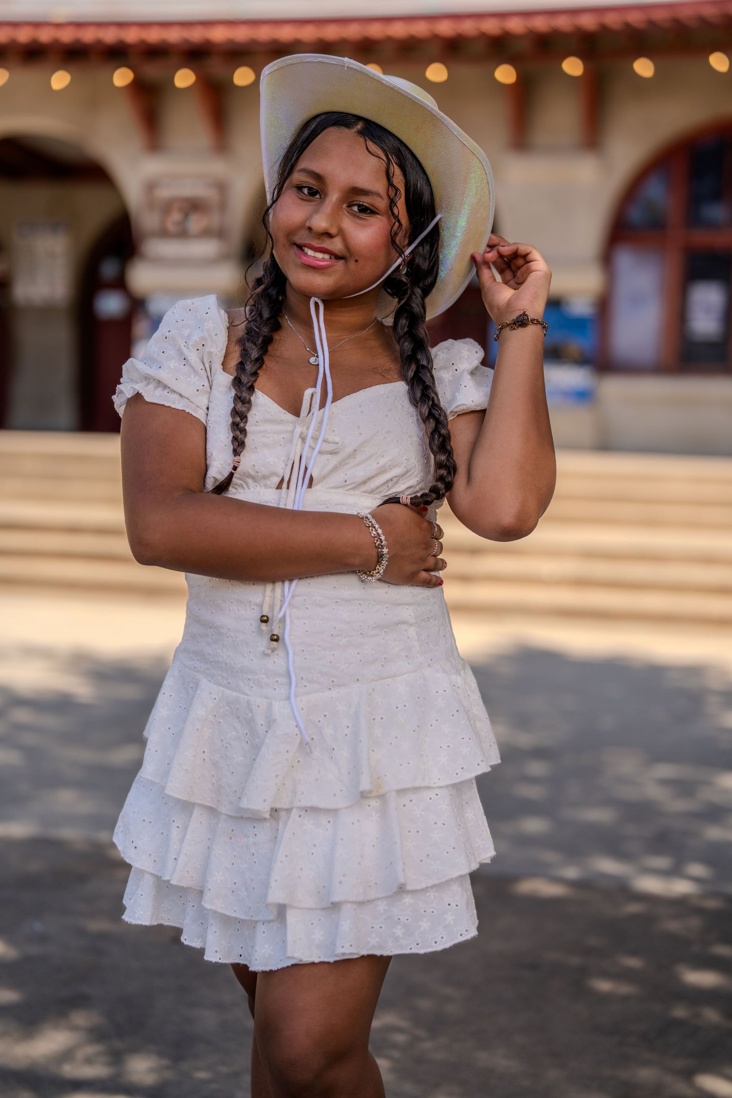 A young dancer in a white ruffled dress stands in a graceful pose with hands raised at an outdoor venue.