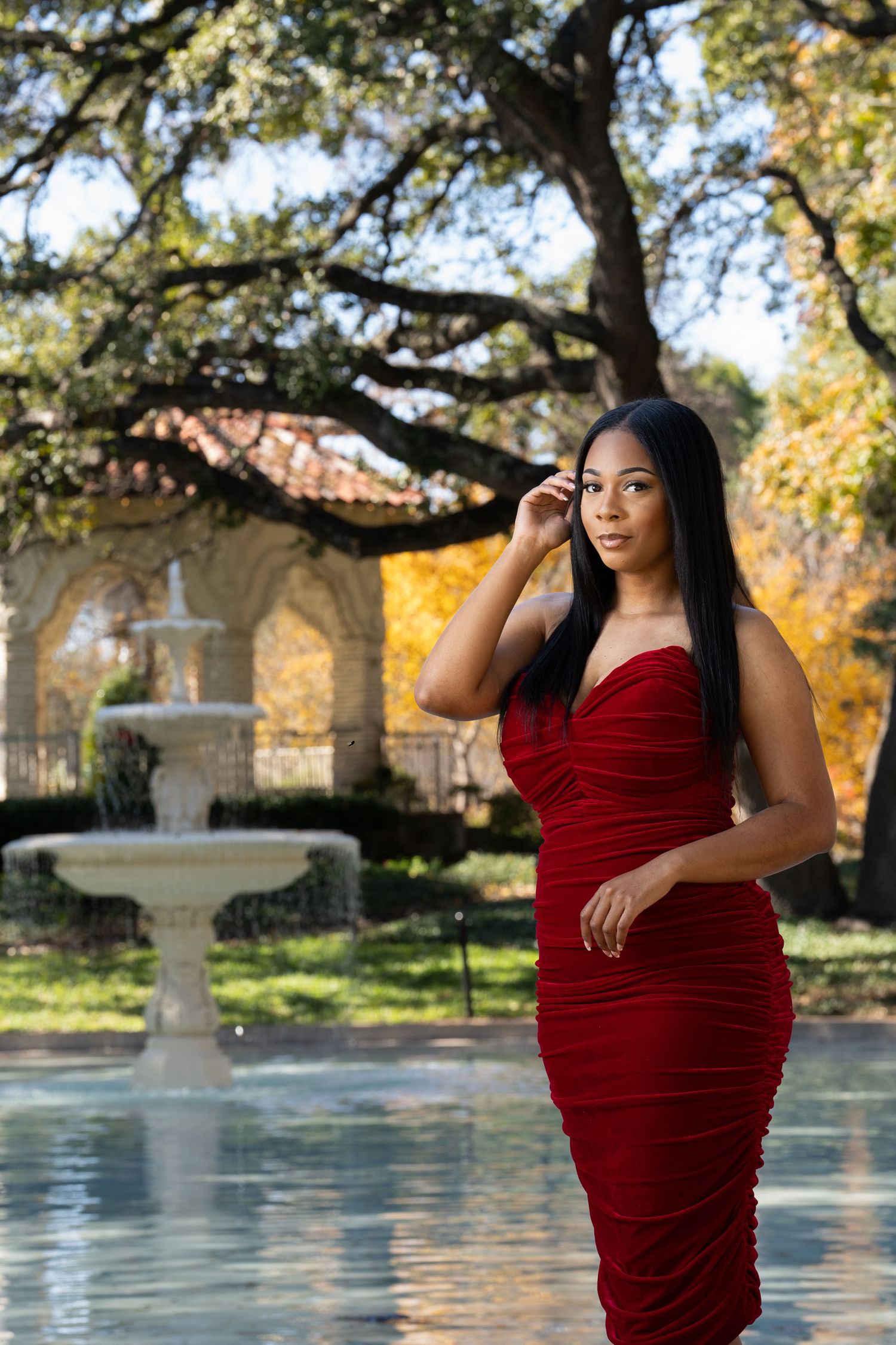 A woman in a deep red velvet dress poses gracefully in an outdoor garden setting with autumn trees and a fountain.