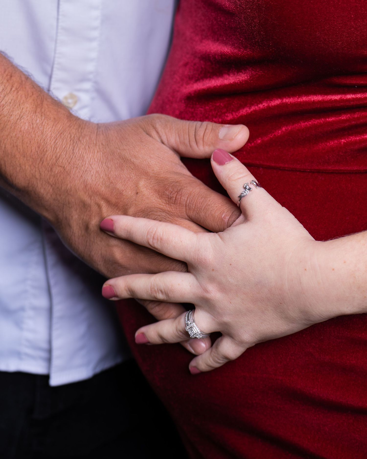 Close-up of couples hands with engagement rings against red velvet dress.