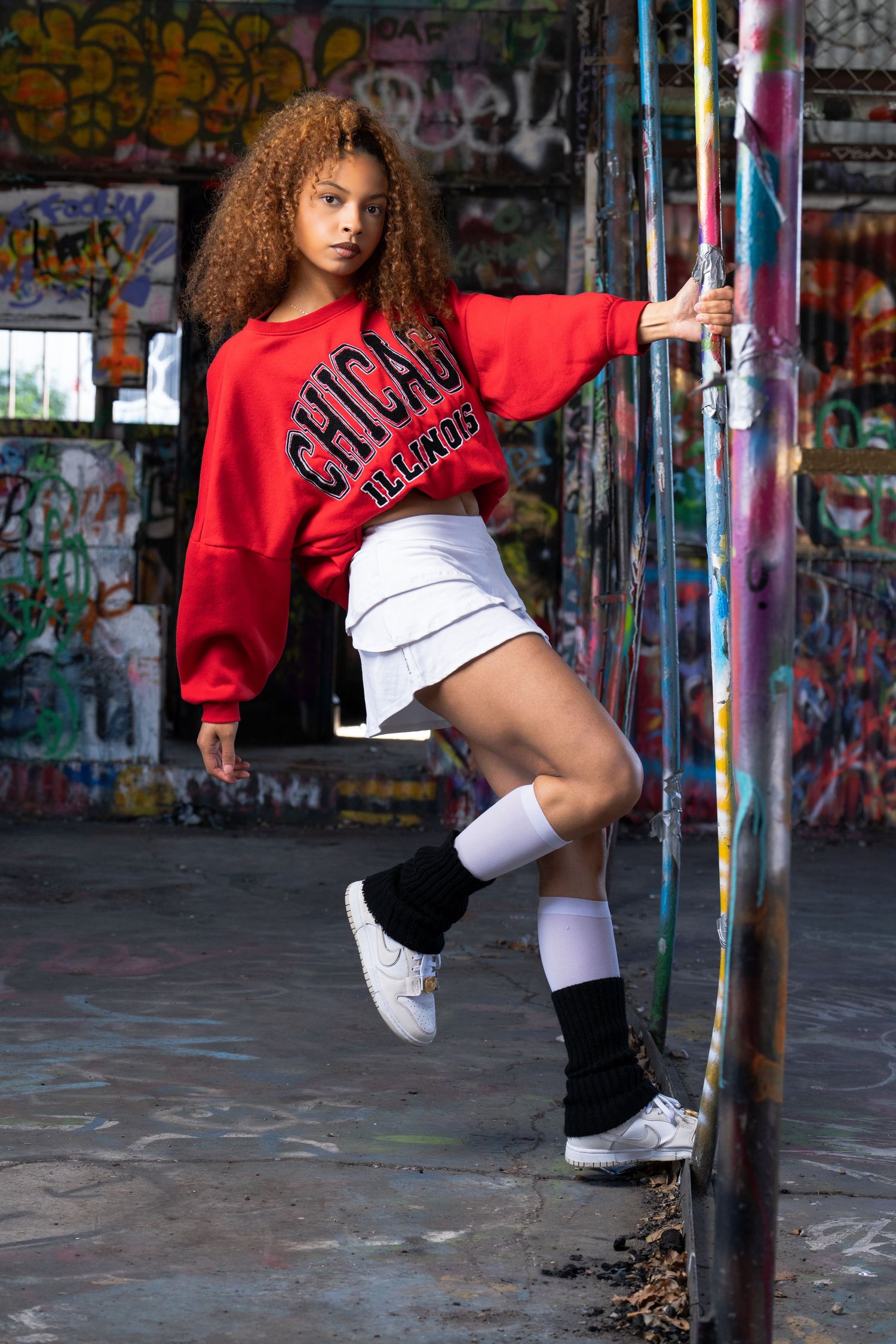 Dancer in red Chicago sweater and white skirt poses against graffiti wall while holding metal pole.