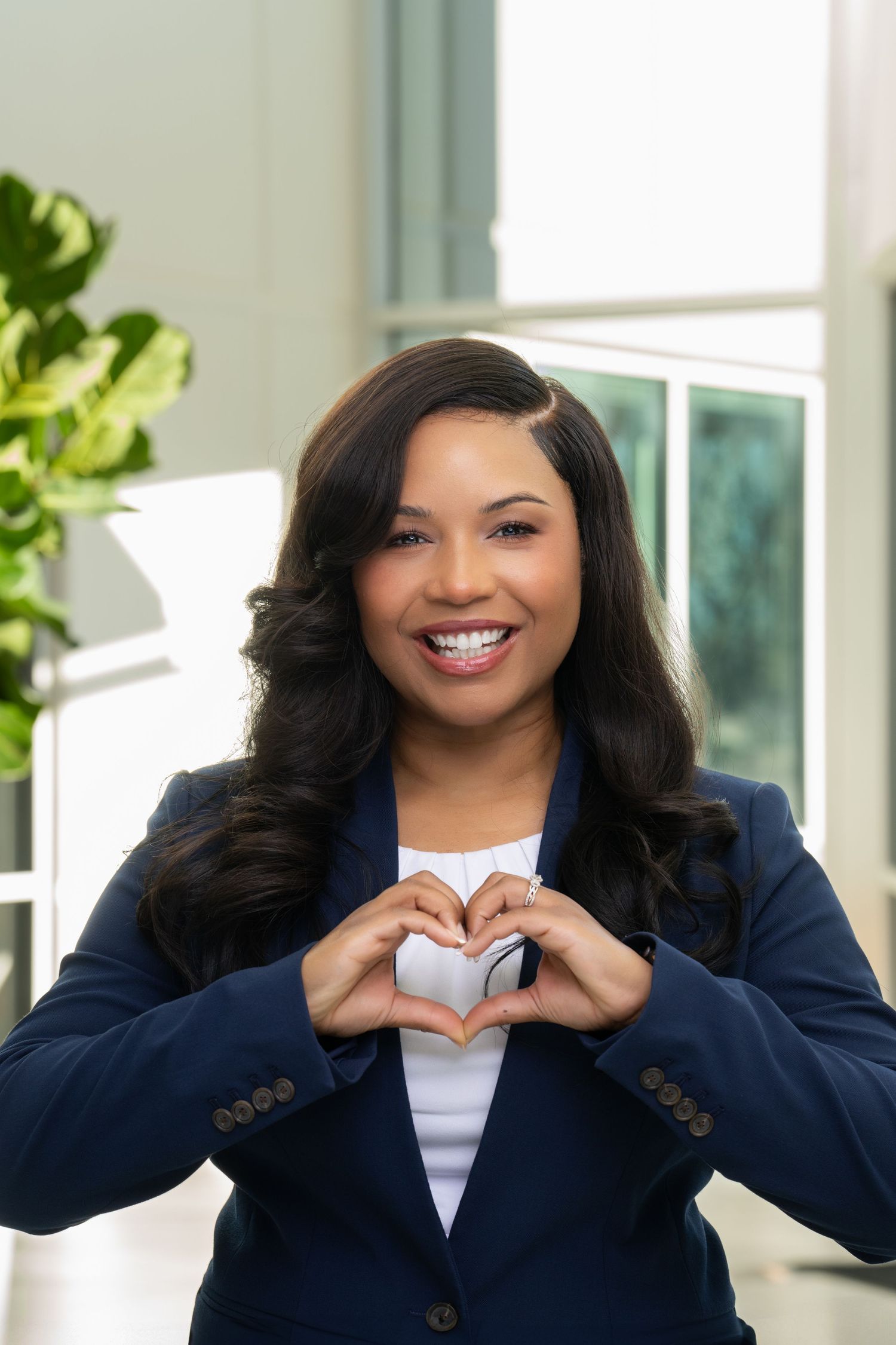Person wearing navy blazer makes heart shape gesture while smiling in bright modern interior.