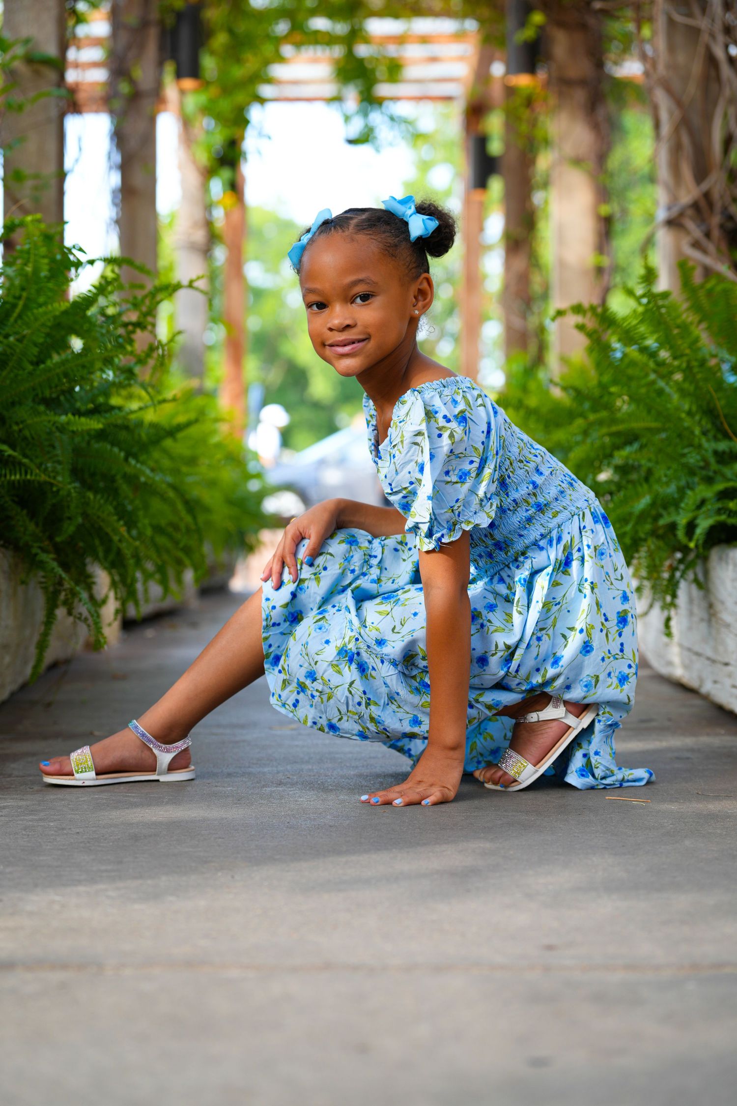 Model in blue floral dress poses playfully in garden pathway lined with ferns.
