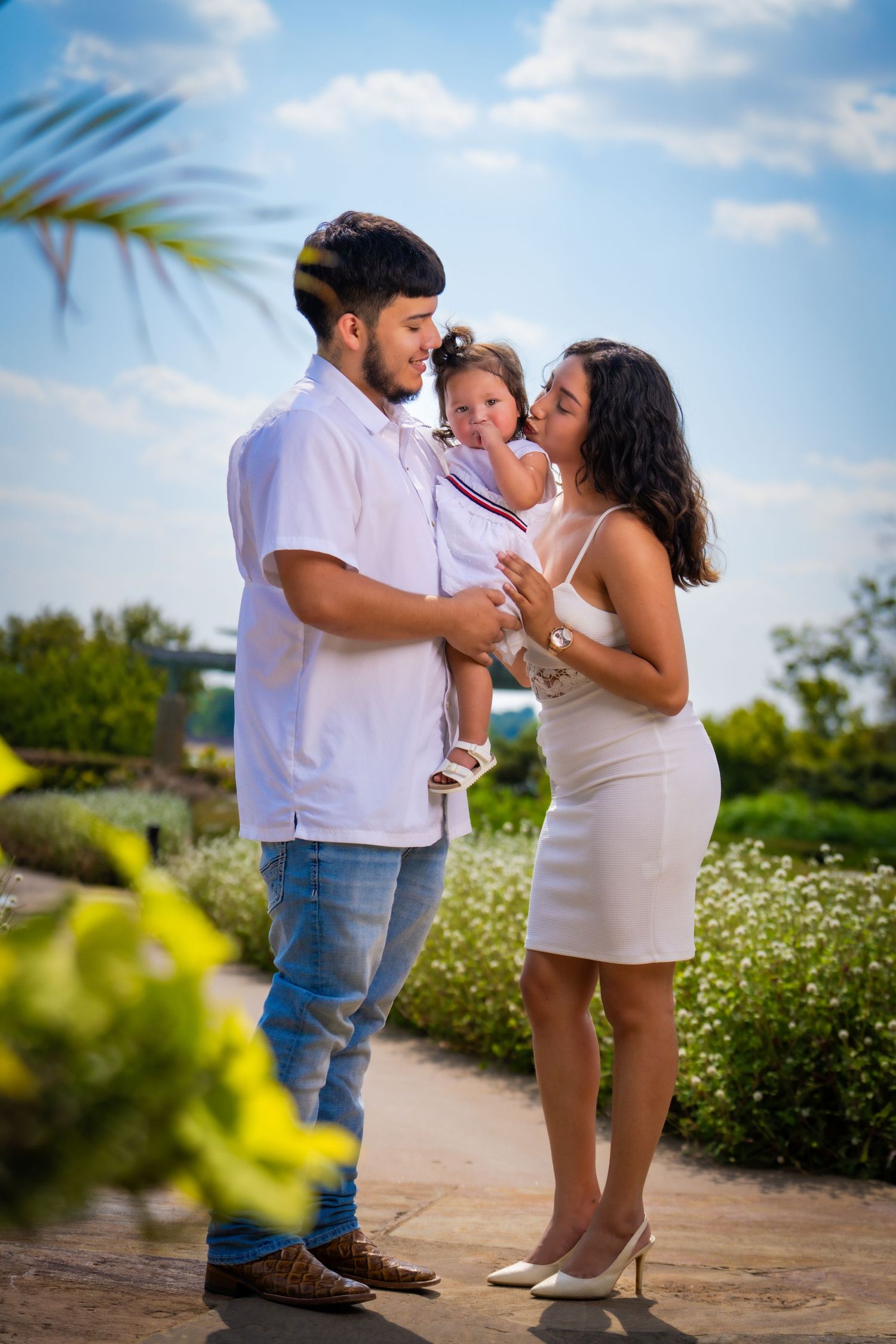 A couple shares a tender moment with their baby in a sunny outdoor garden setting.