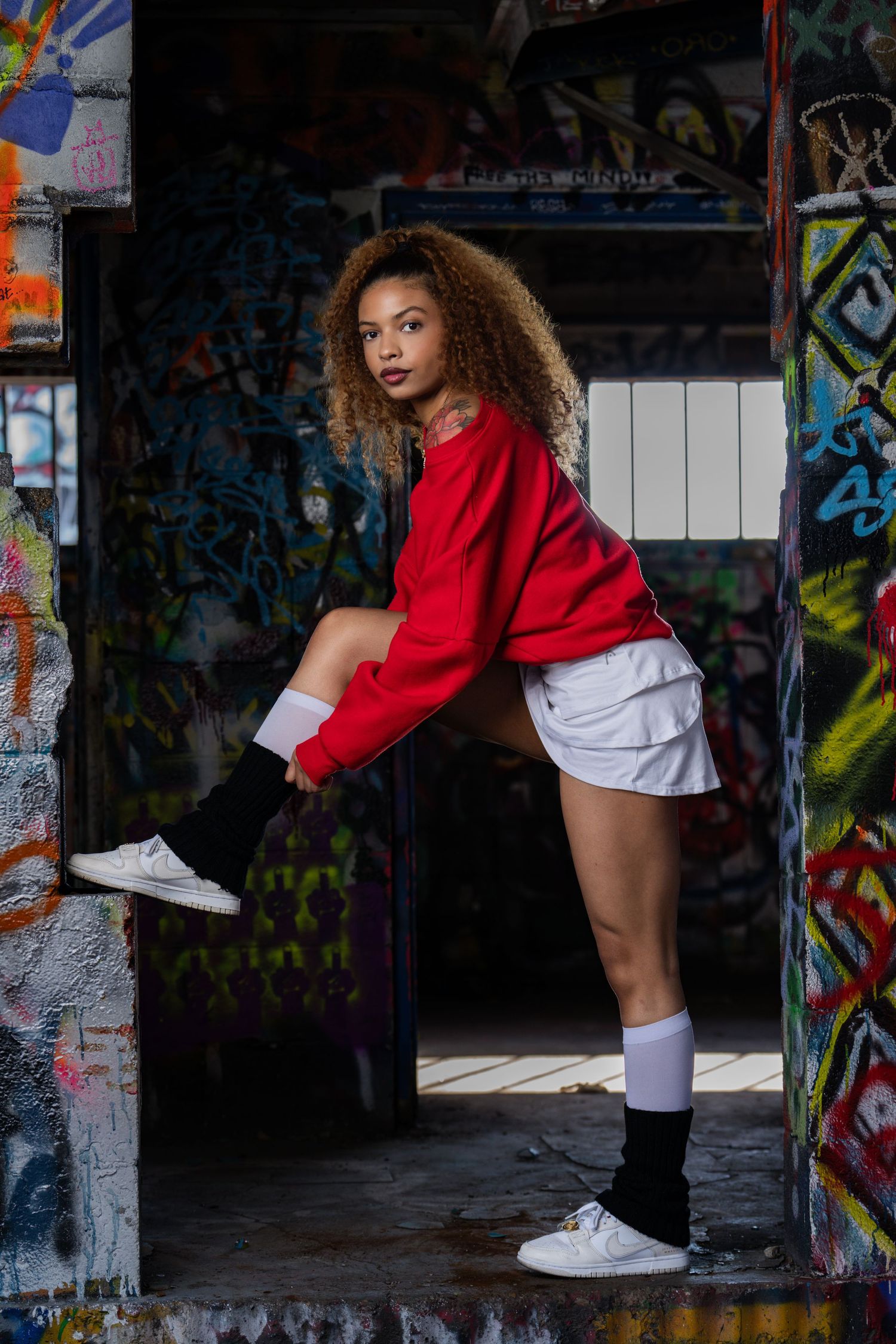 Fashion model in red sweater and white shorts poses against graffiti wall in urban setting.