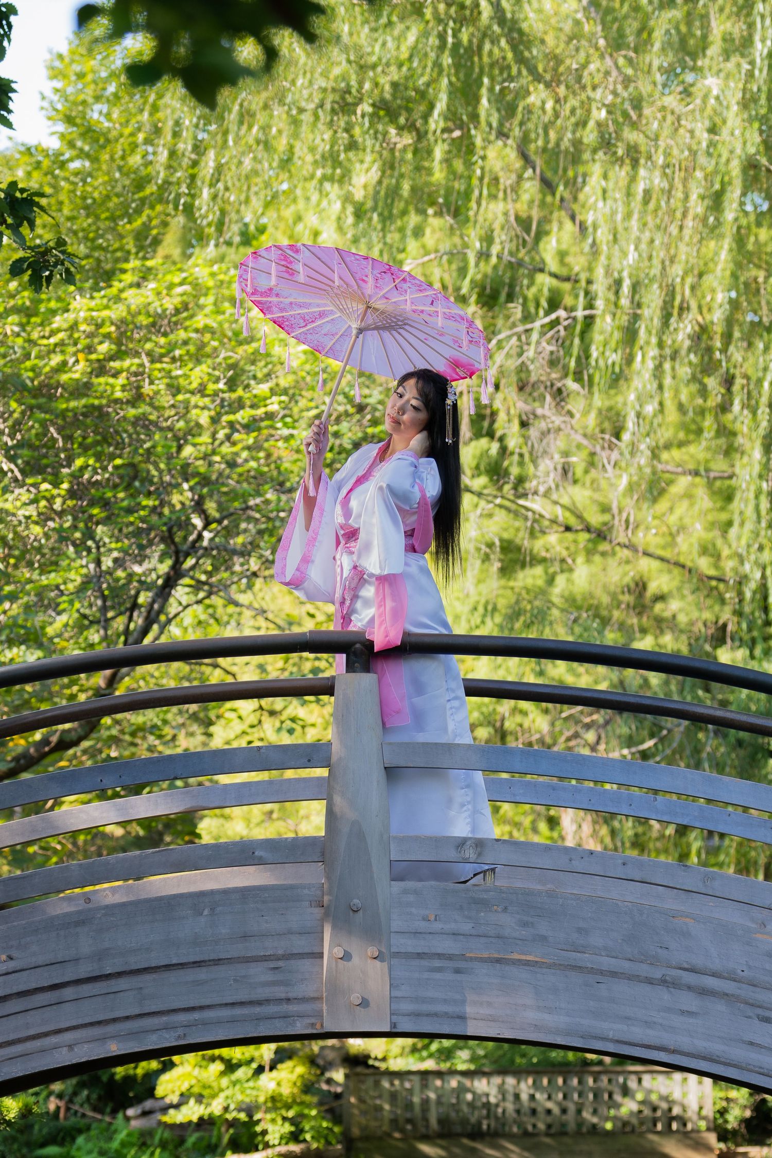 A figure in a white dress and pink parasol poses gracefully on a curved wooden bridge in a lush garden setting.