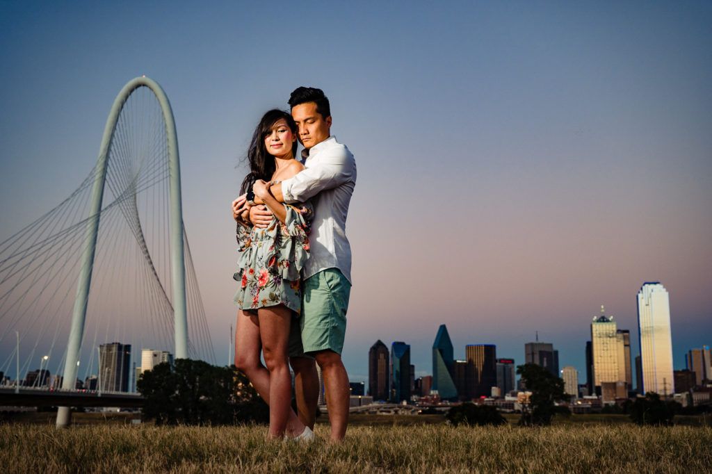 A couple embraces on a grassy hill at sunset with the Dallas skyline and Margaret Hunt Hill Bridge in the background.