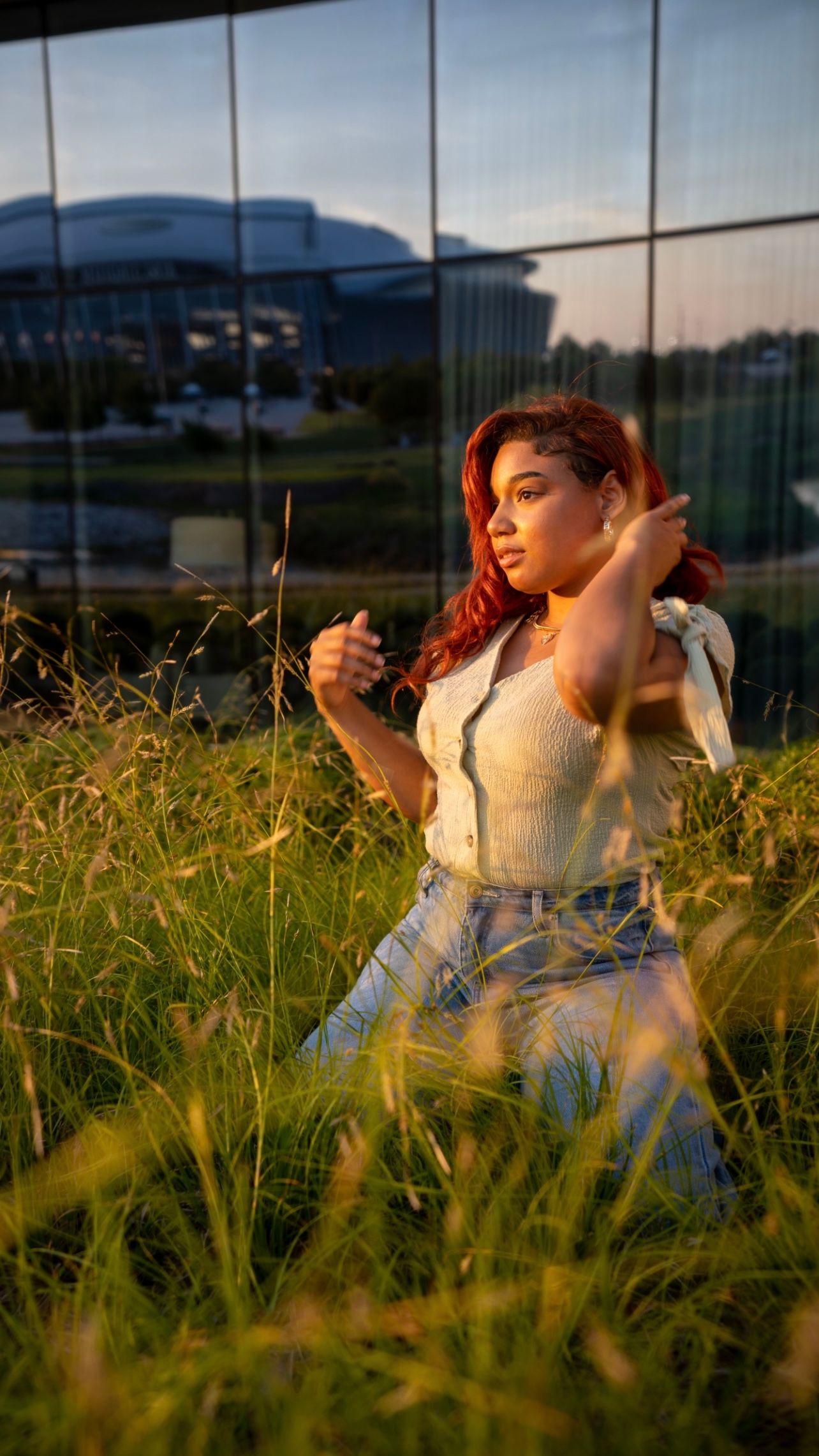 A person sits in tall grass at sunset near a modern building with reflective glass windows. Professional  Birthday photos in Dallas Texas