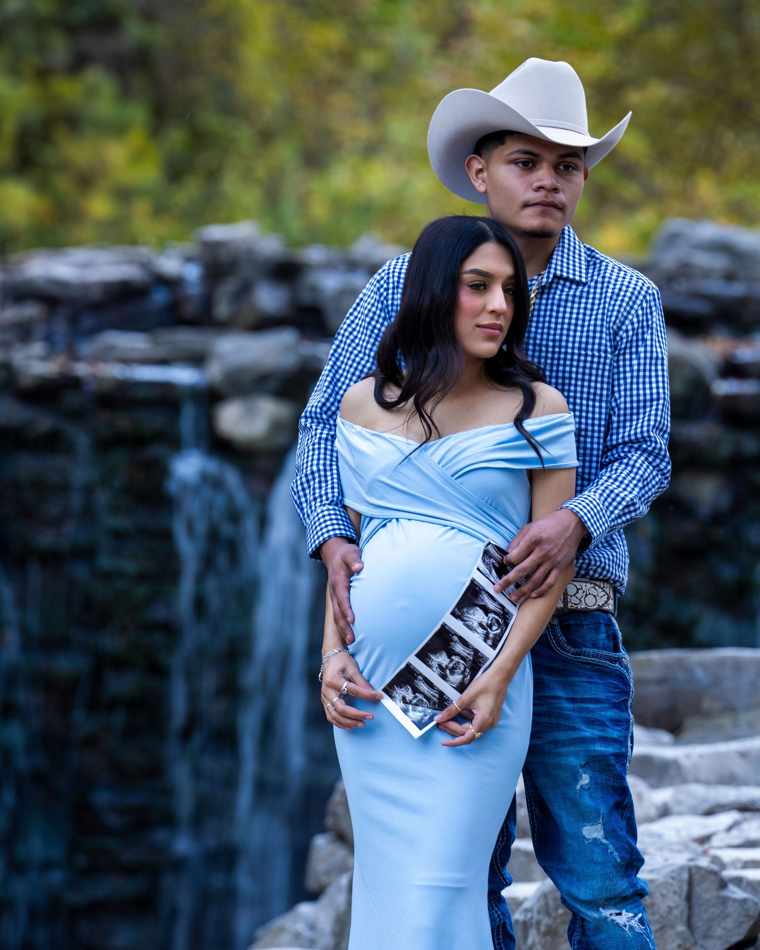 Maternity photo of couple in western wear standing by waterfall holding ultrasound image.