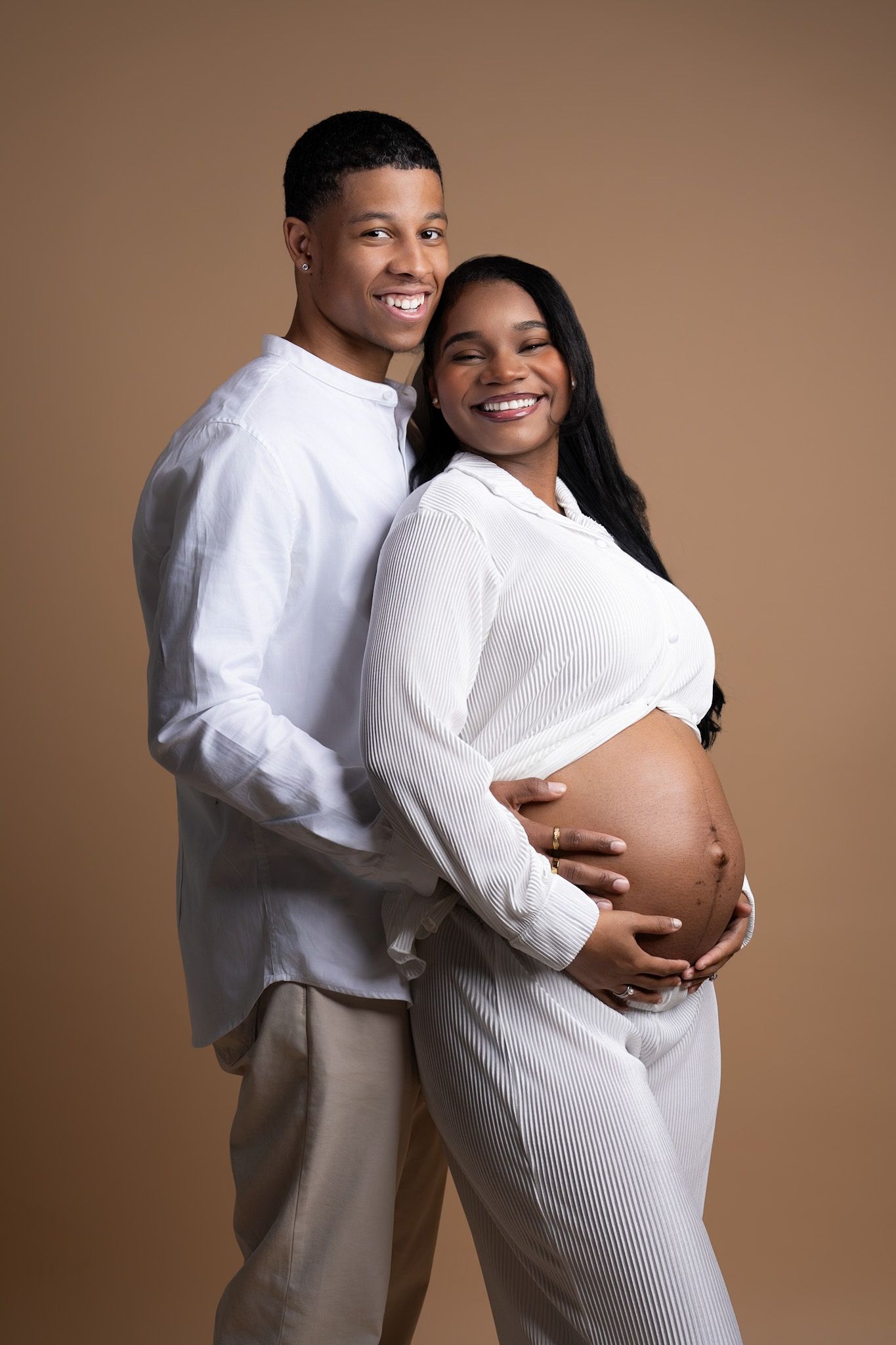 Happy couple poses for maternity photo in matching white outfits against warm brown background.