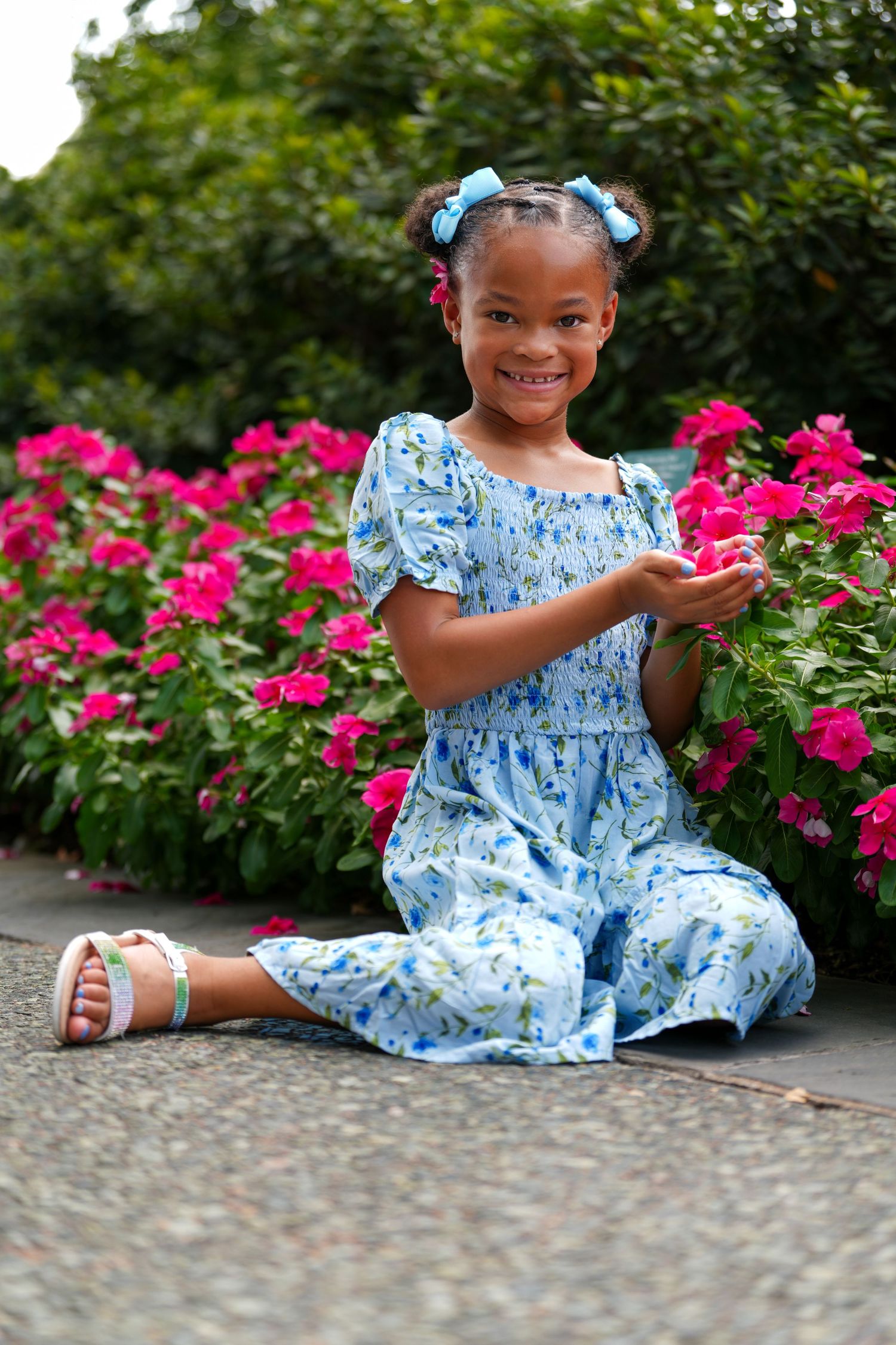 Young girl in blue floral dress sits by vibrant pink garden flowers on a stone path in a scenic outdoor setting.