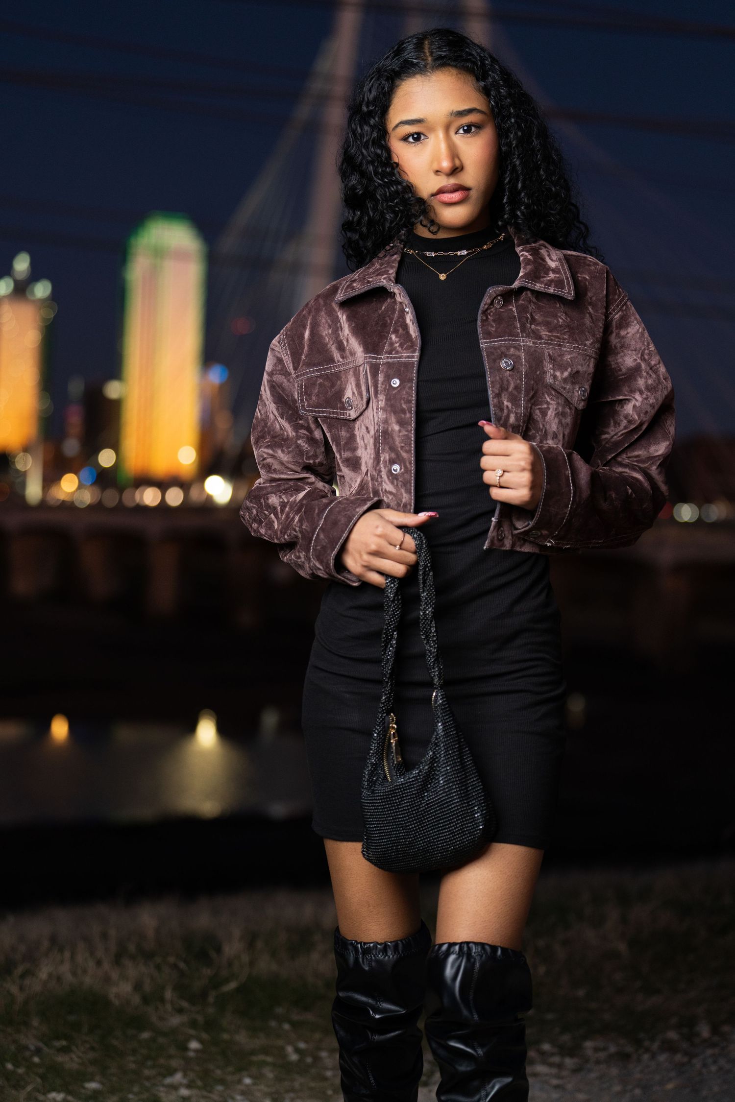Fashion model wearing brown leather jacket and black boots poses at night against illuminated city skyline.