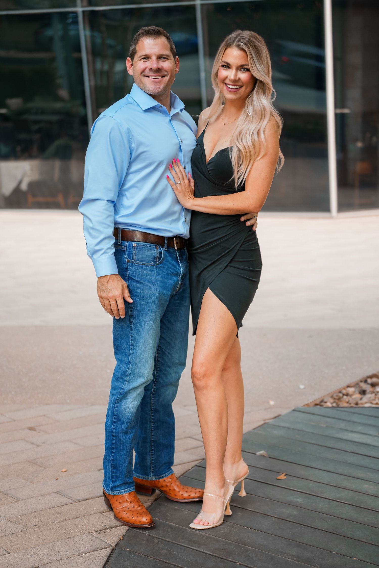 A couple poses together outdoors, with him in a light blue shirt and jeans and her in an elegant black dress.