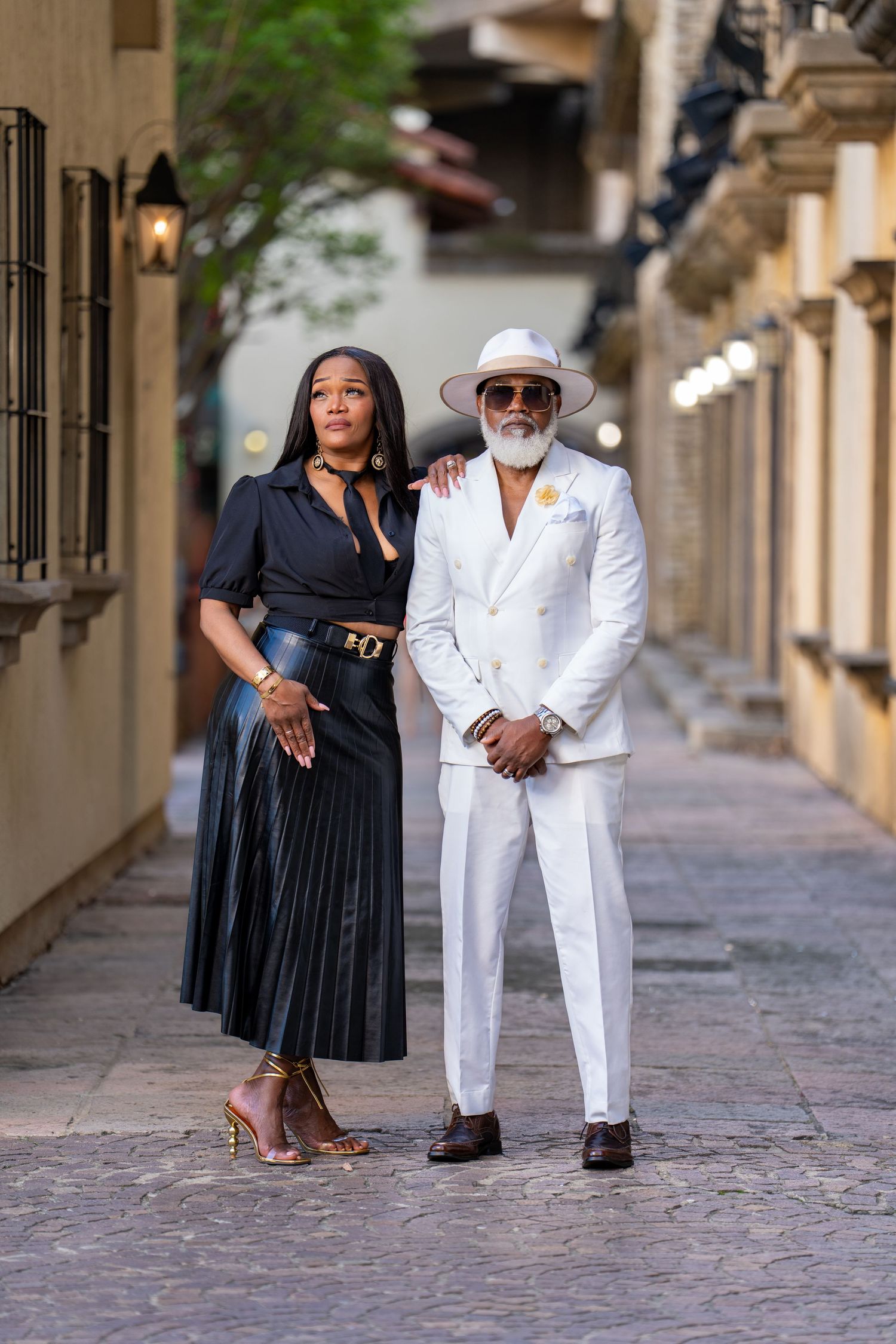 Stylish couple poses in a cobblestone alley wearing contrasting black and white formal outfits.