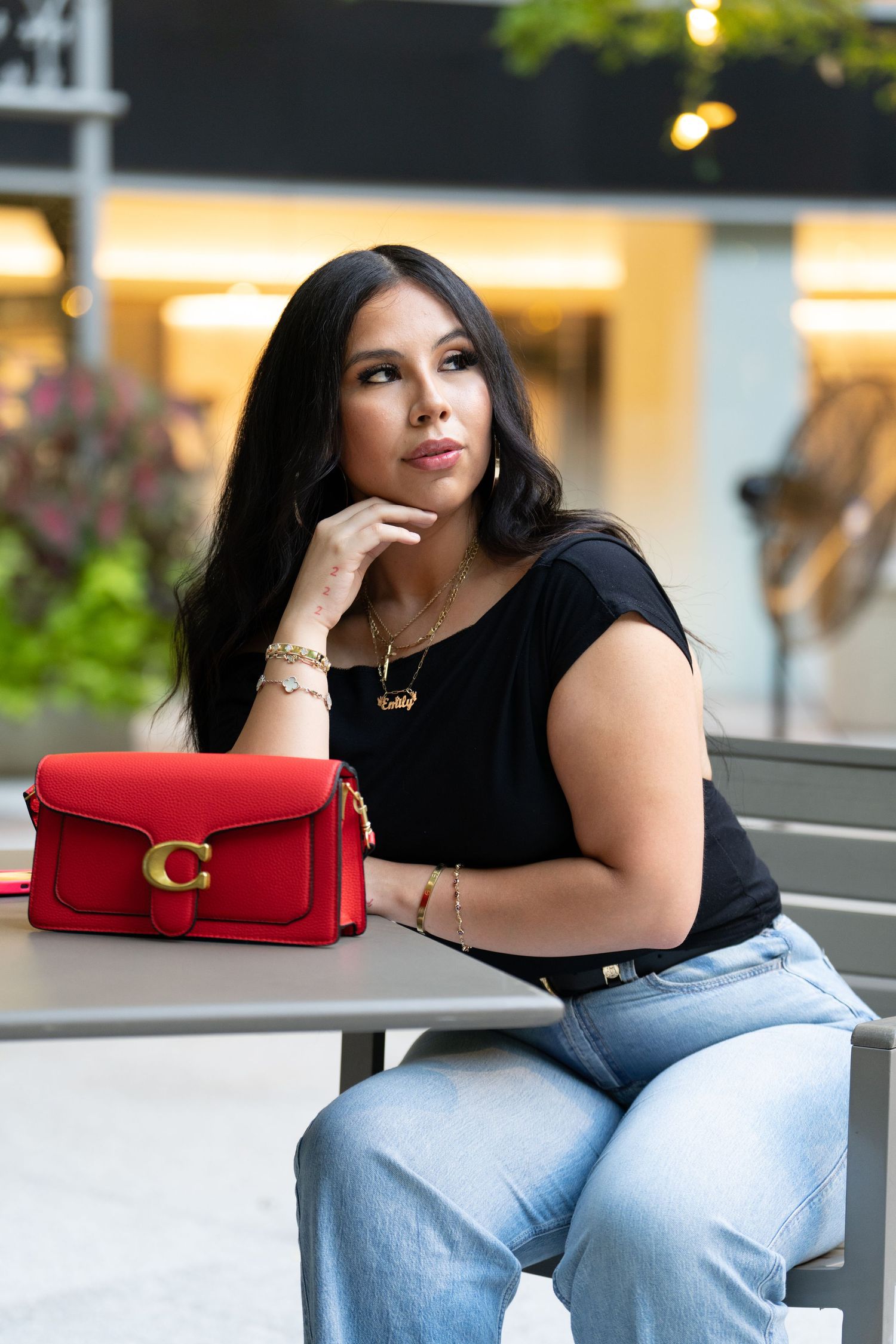 Model wearing blue jeans and black top sits casually with red designer handbag in outdoor fashion setting.