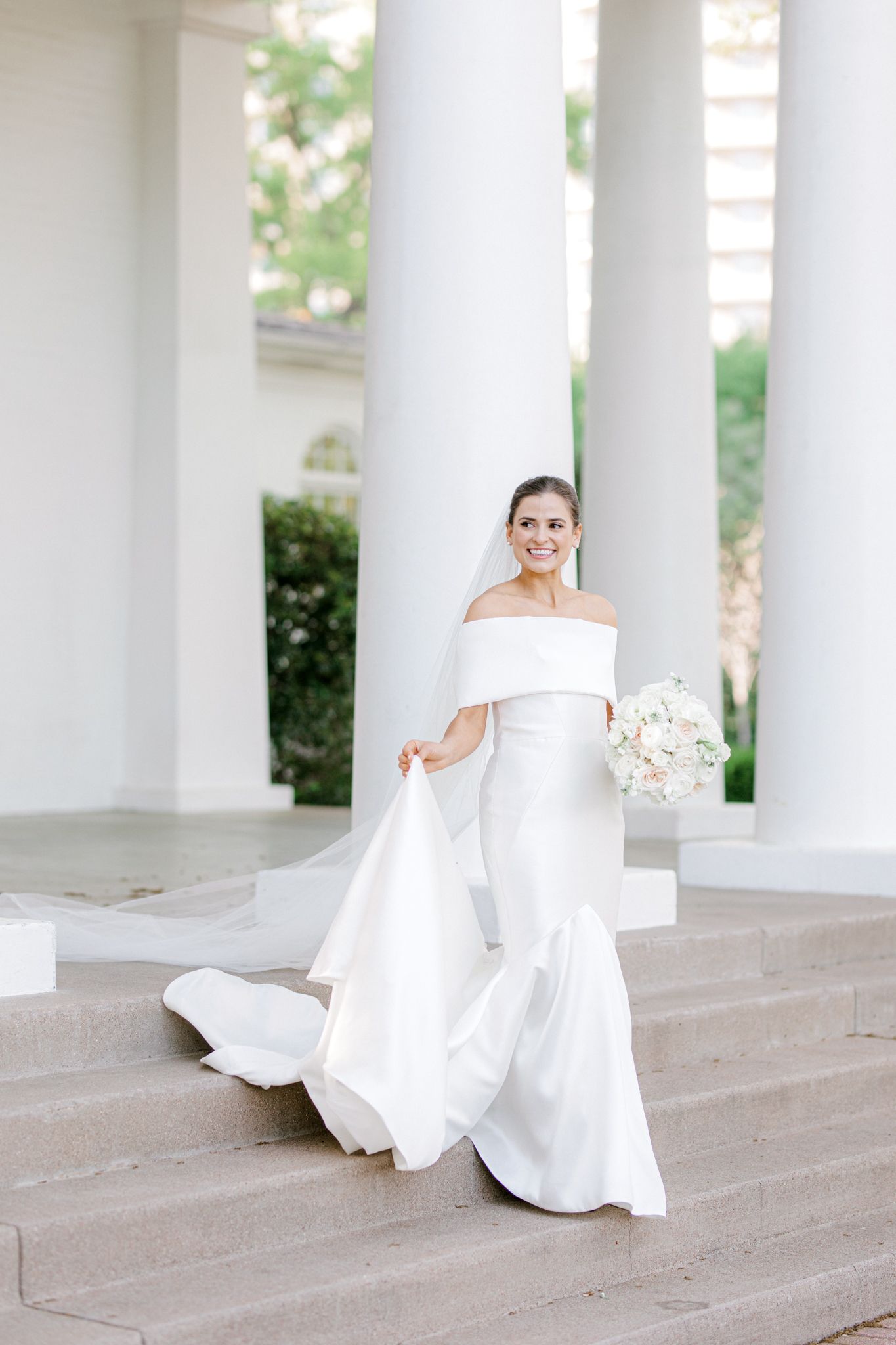 Elegant bride in modern off-shoulder white wedding gown with long train stands gracefully between classic white columns.