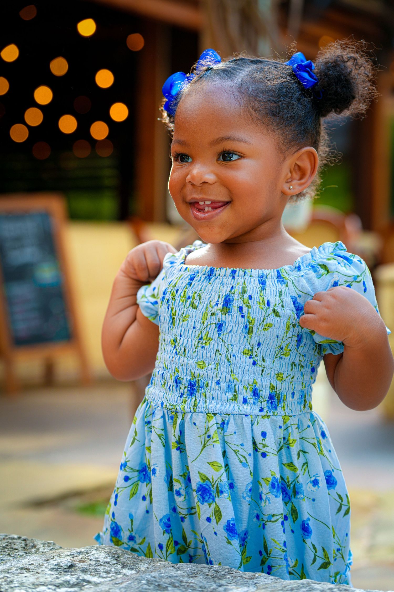 Small child in blue floral dress with matching hair bows against bokeh background.