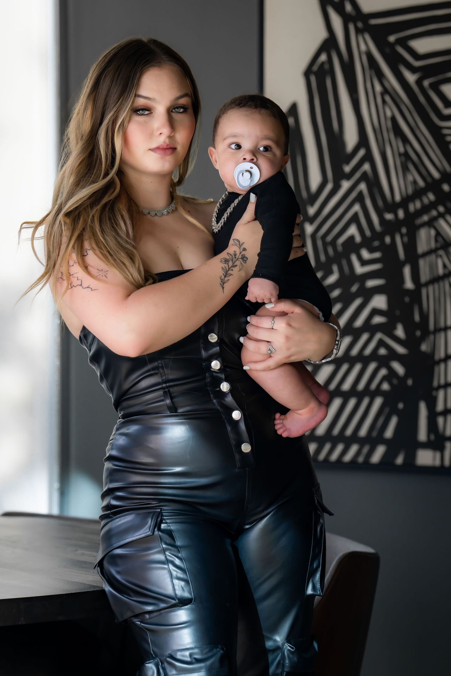A parent in a black leather outfit holds a baby wearing dark clothing against a geometric patterned wall.