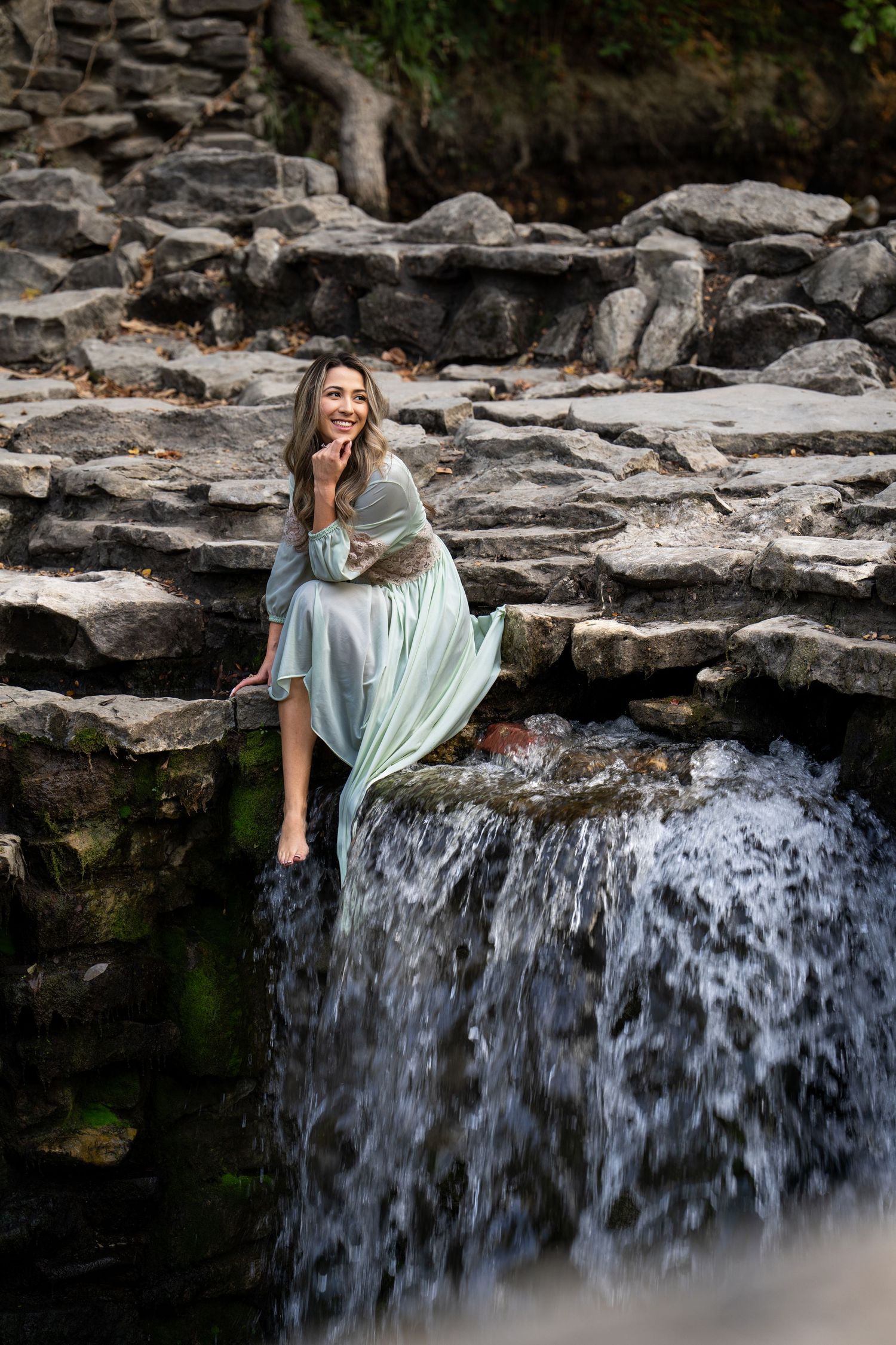 Person in flowing mint dress poses gracefully on rocky waterfall ledge in nature setting.