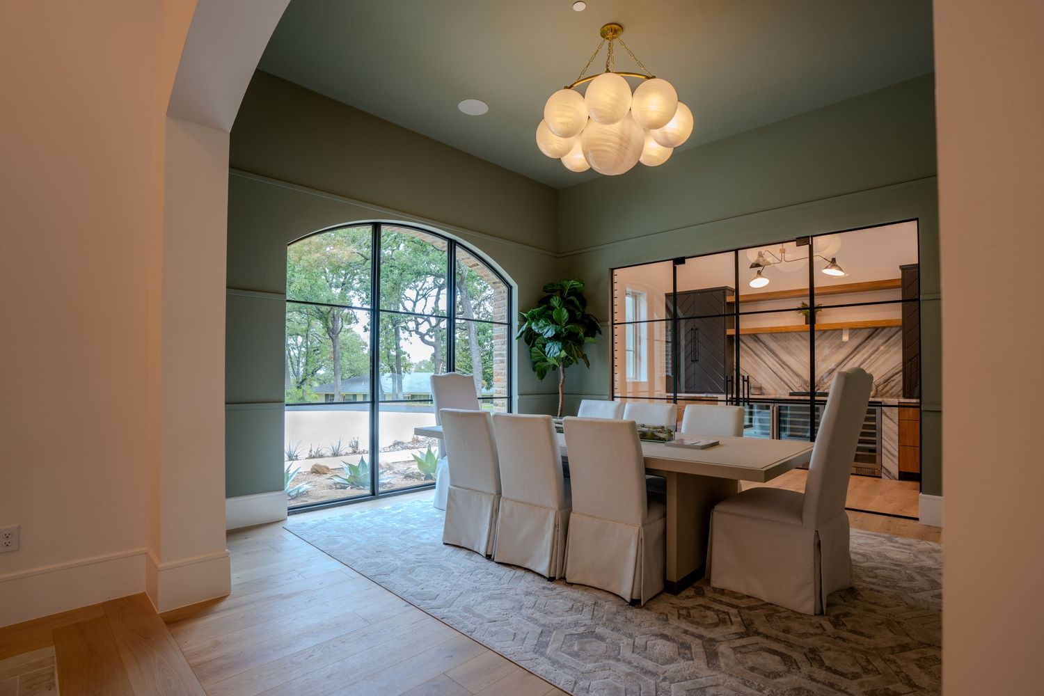 Elegant dining room with arched windows, chandelier and upholstered chairs.