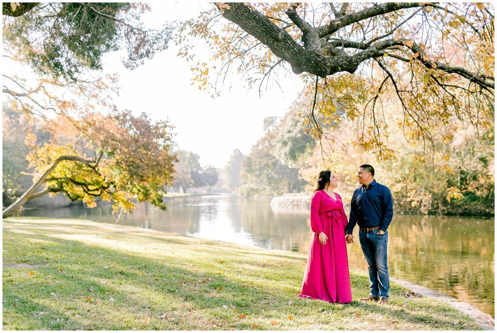 Two people stand near a serene lake surrounded by autumn trees with golden leaves at sunset.
