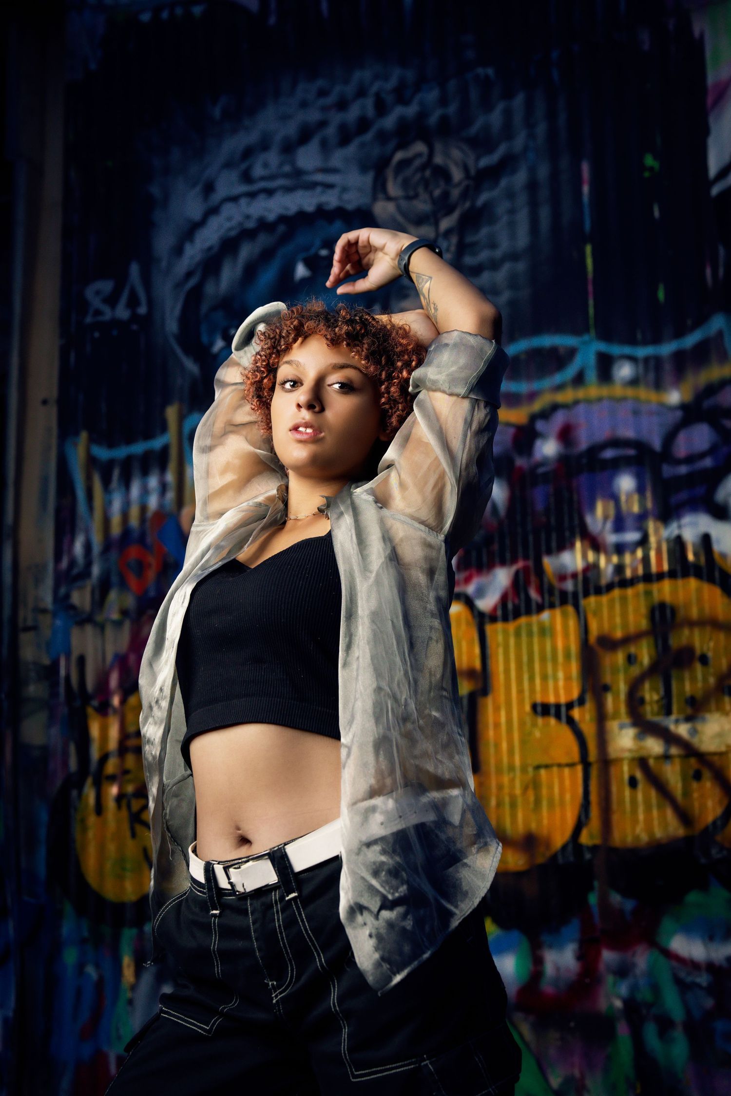 Fashionable model in black crop top and sheer jacket poses against colorful graffiti wall.