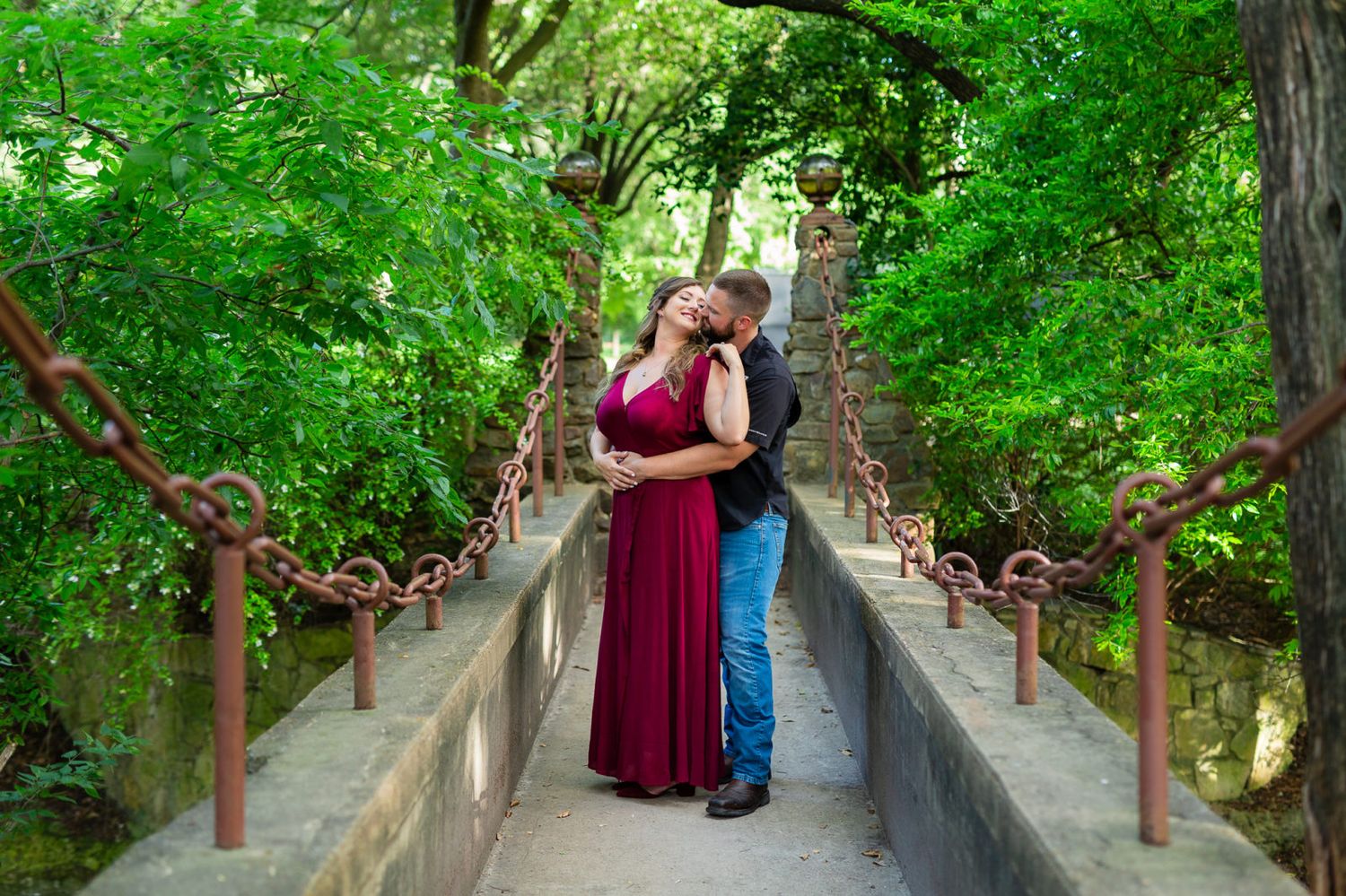 A romantic moment shared on a wooden bridge surrounded by lush green foliage in a natural setting.