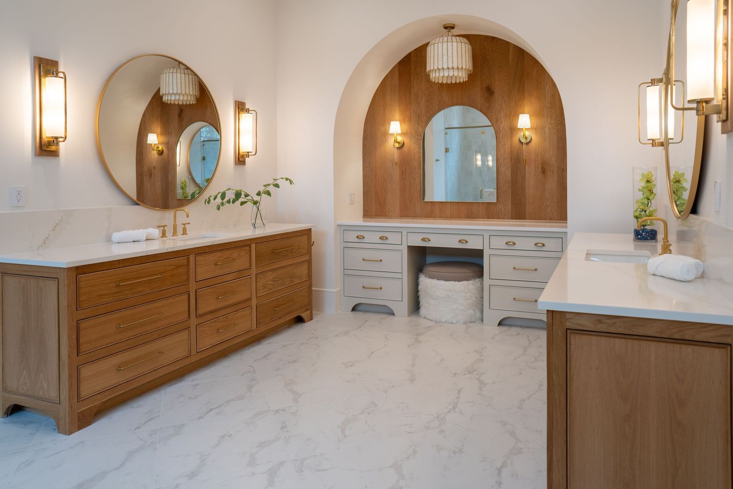 Modern bathroom featuring wood vanities, arched mirrors, and marble flooring with warm lighting.