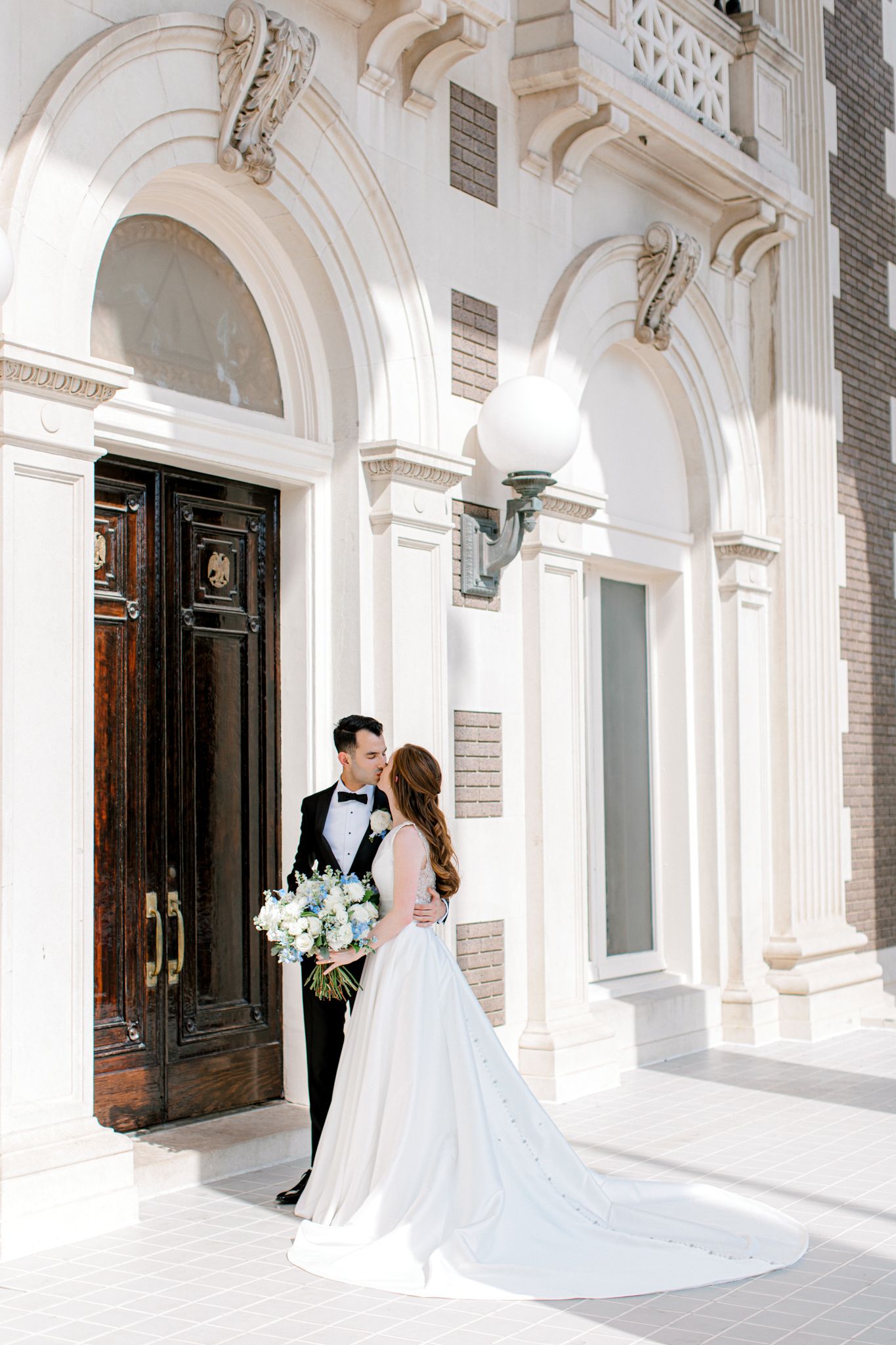 Couple in formal wedding attire standing in front of ornate white stone archways with classical architectural details.