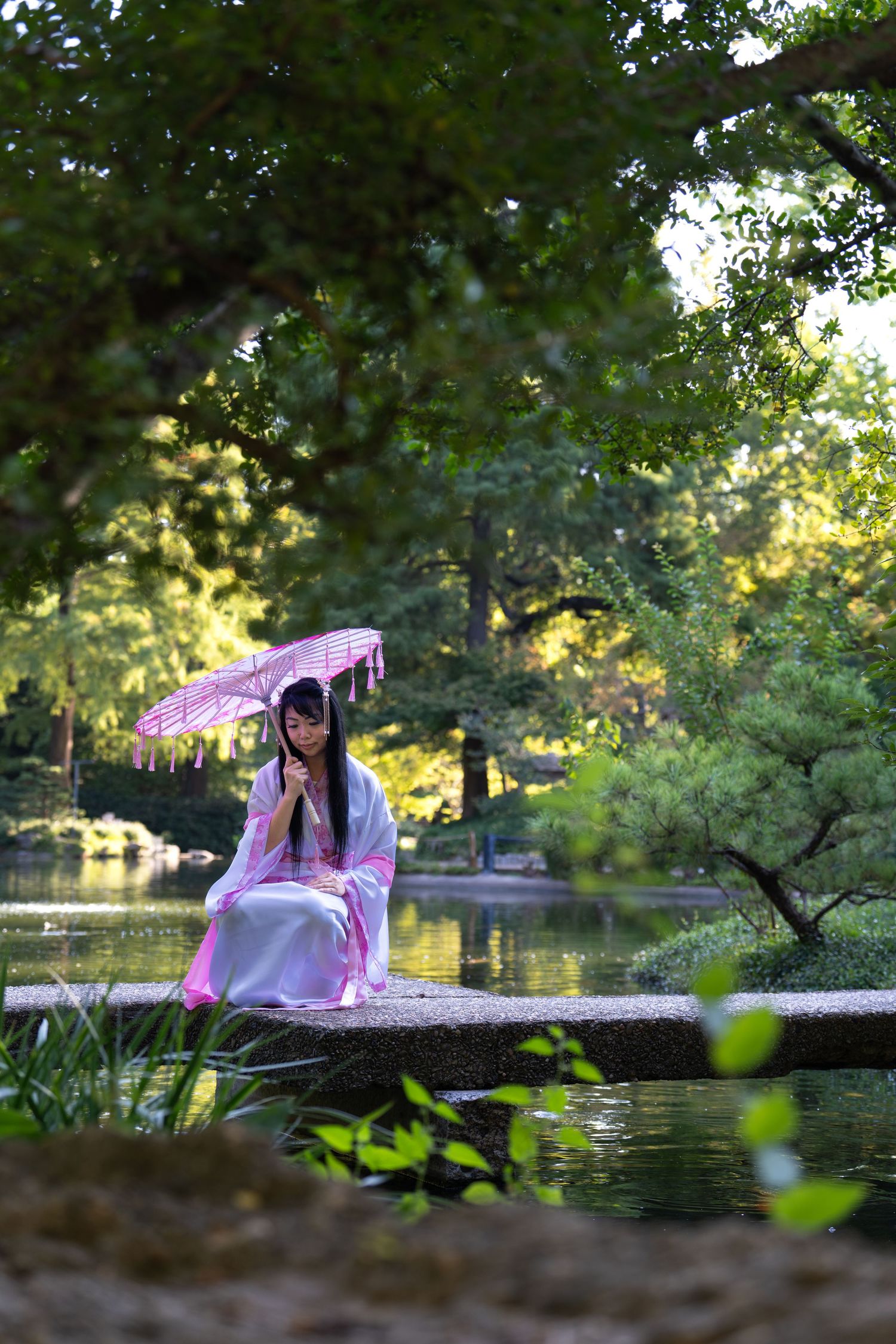 A person in pink clothing lies on their stomach near a tranquil pond in a lush park setting.