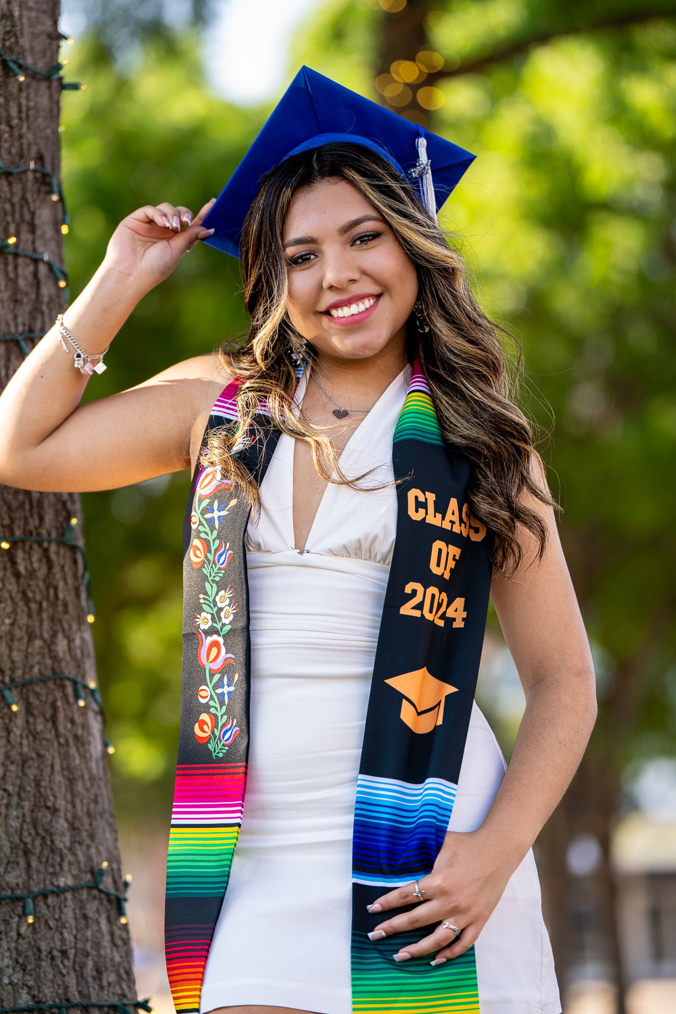 Graduate wearing white dress and decorated stoles poses confidently by tree.