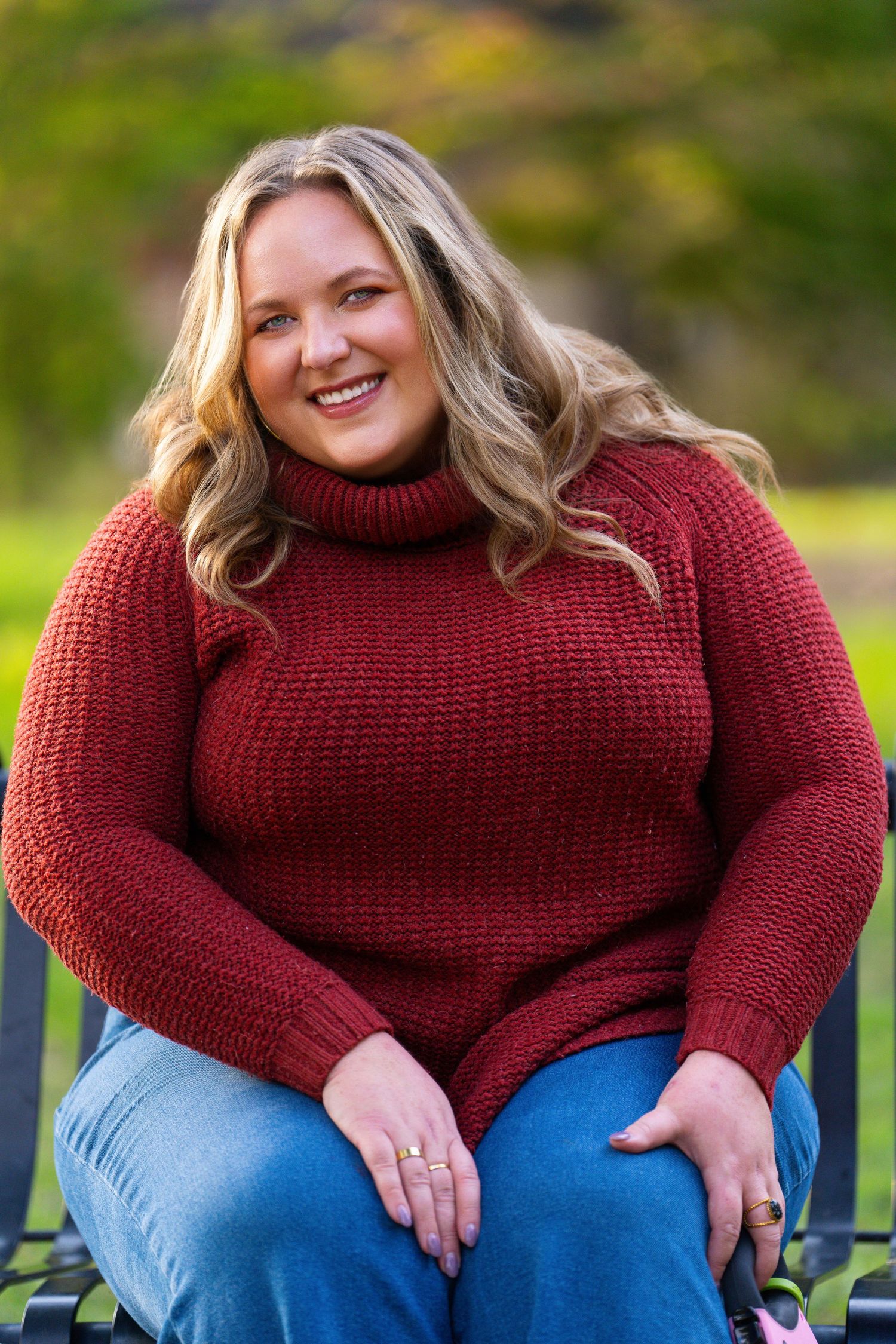 A casual outdoor portrait of someone wearing a burgundy turtleneck sweater and blue jeans seated on a bench.