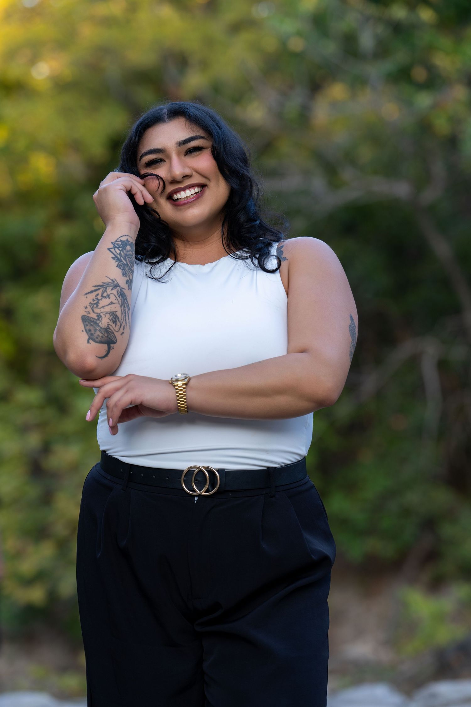 A curvy person wearing a white top and black pants poses outdoors with a joyful expression and visible arm tattoo.
