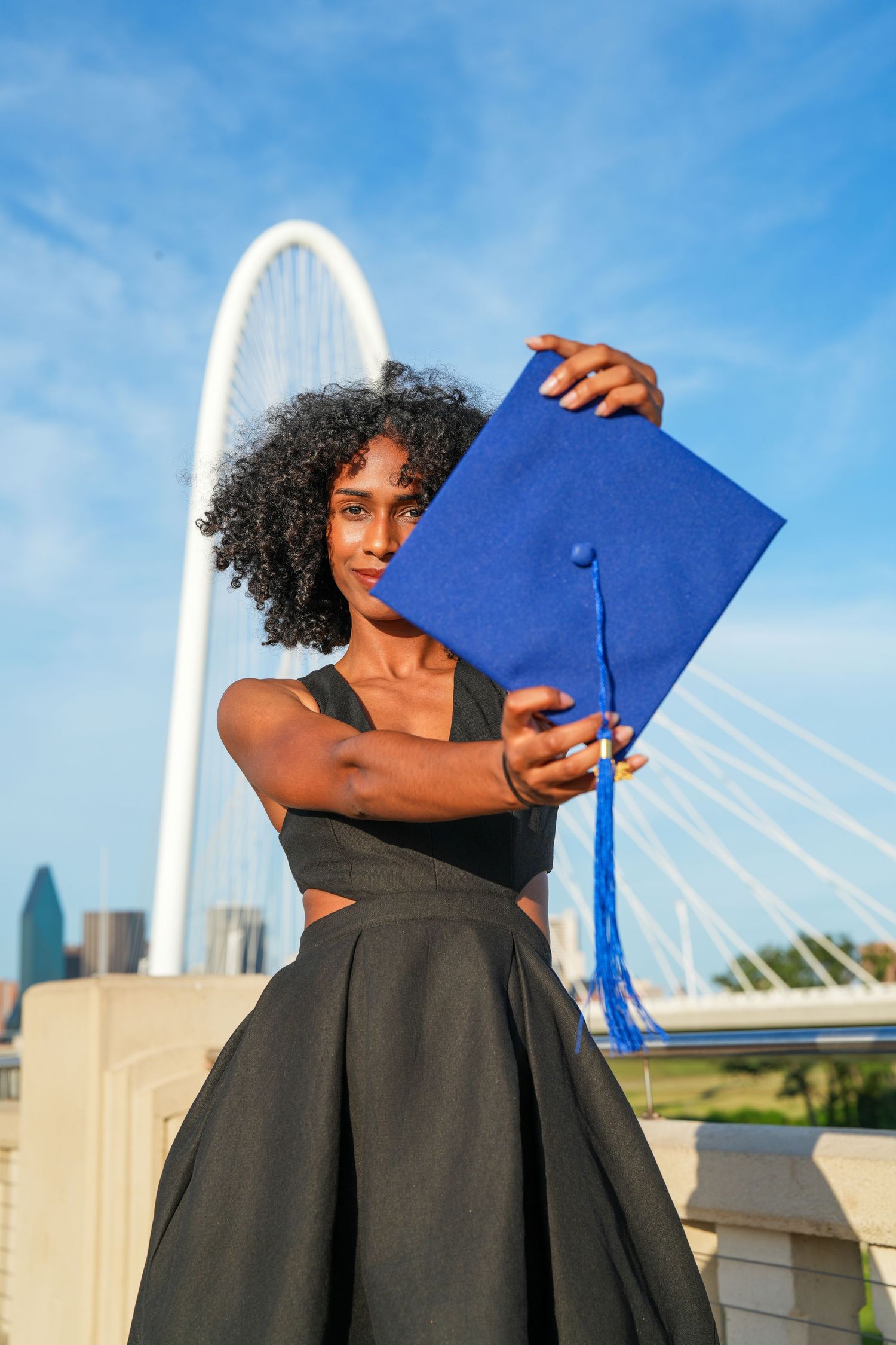 Graduate holds up blue diploma cover in front of the iconic Margaret Hunt Hill Bridge in Dallas.
