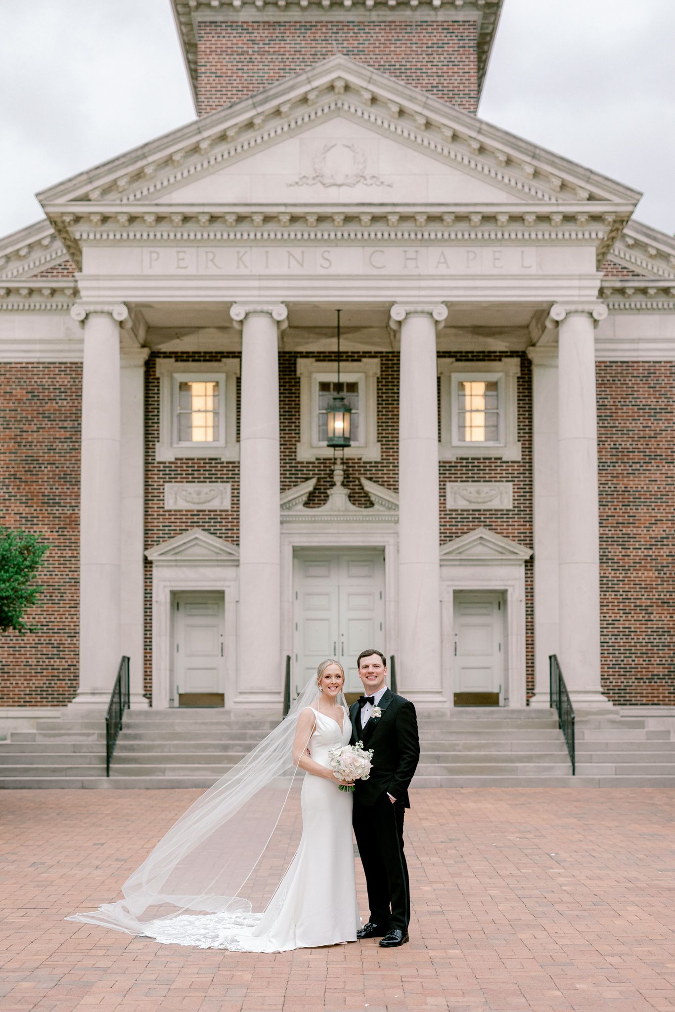 Classic brick and stone church building with white columns and triangular pediment serves as backdrop for elegant wedding photo.
