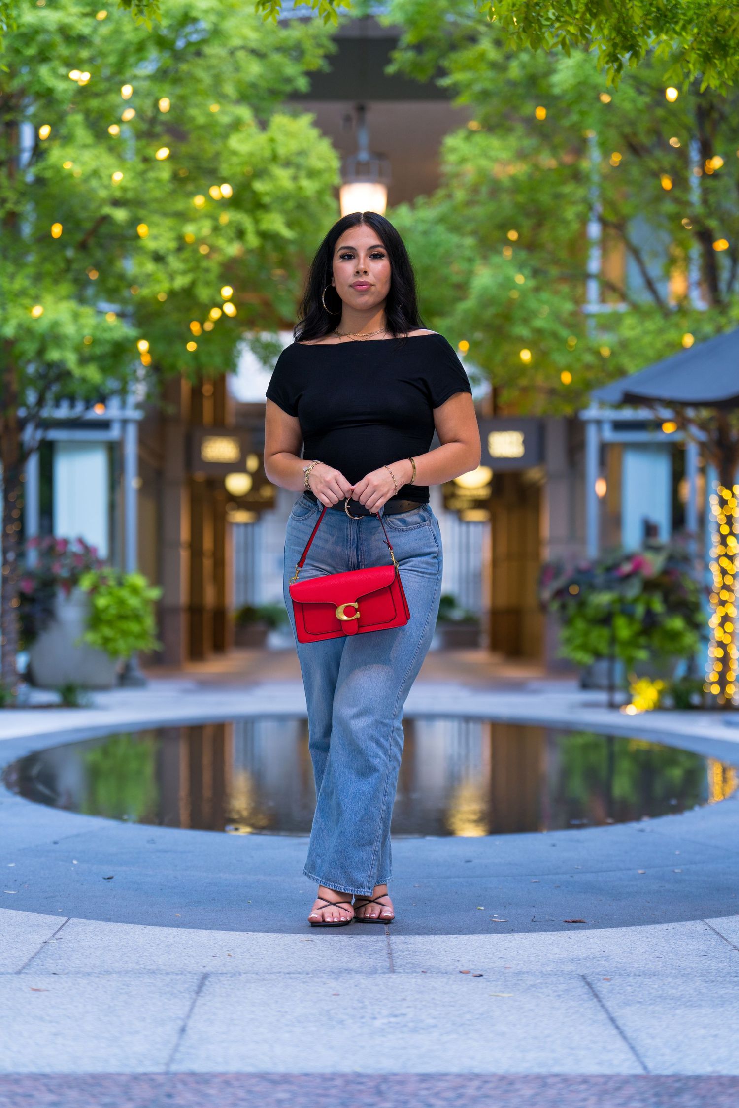 Fashion portrait near reflecting pool with string lights and red designer handbag.