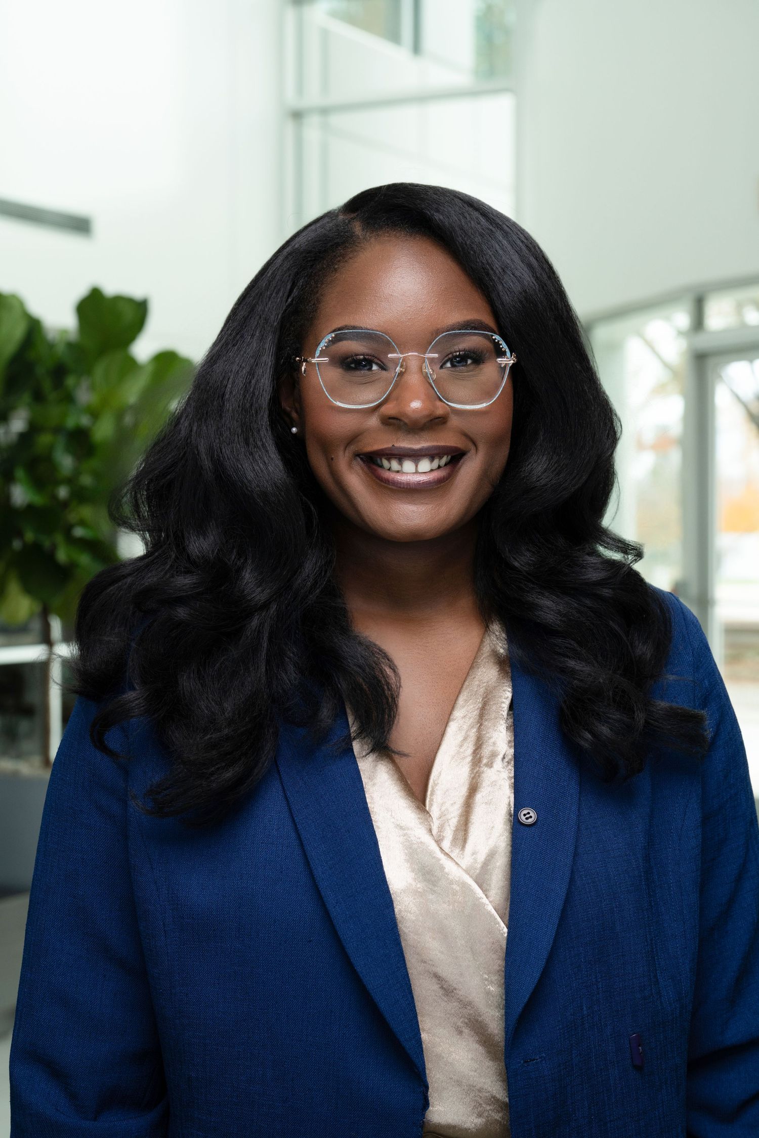 Smiling professional in navy blazer and glasses with styled hair in bright office setting.