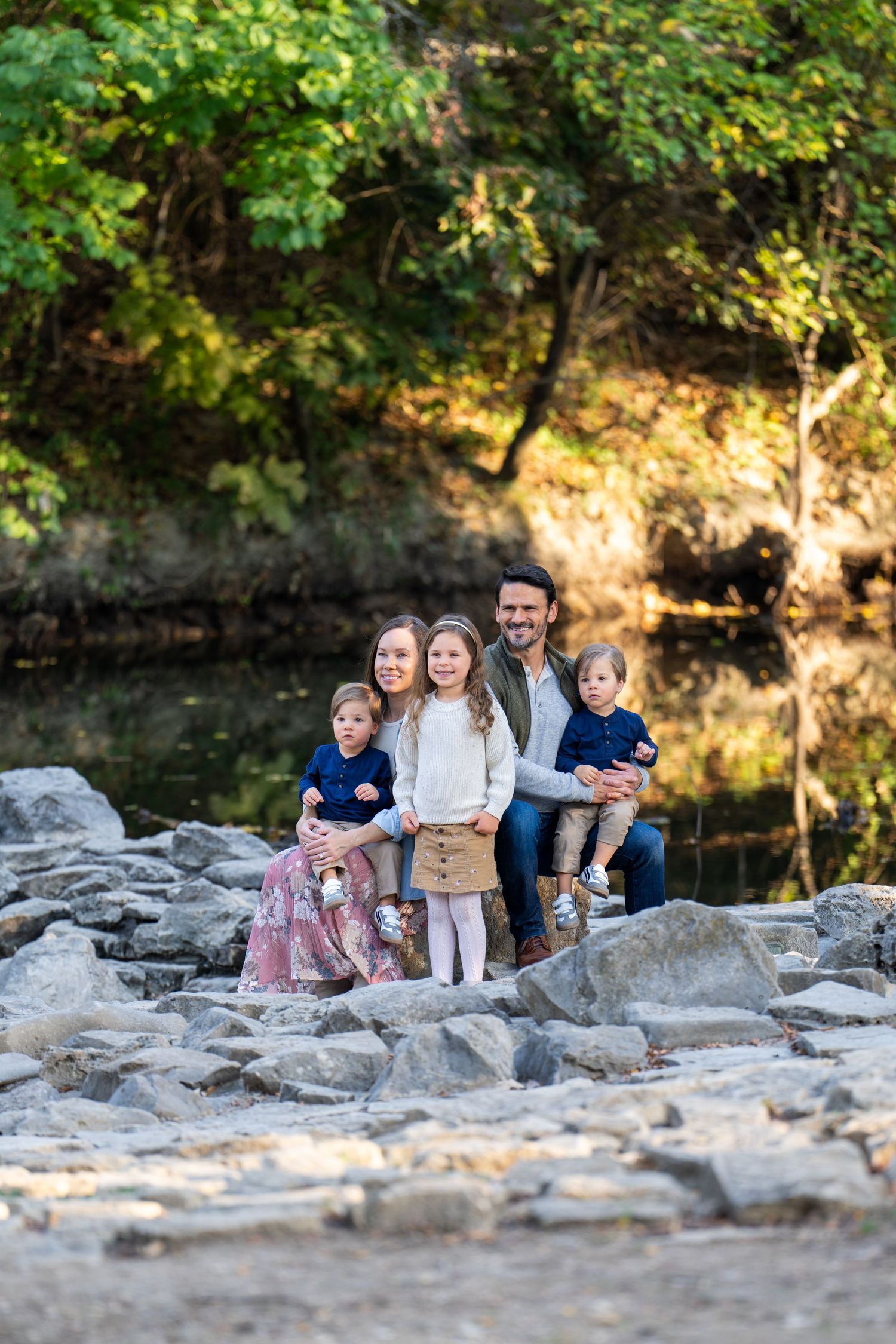 A group sits together on rocky terrain beside a peaceful stream with sunlit trees in the background.