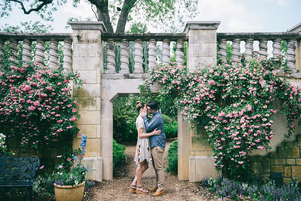 A romantic couple walks together in a garden path surrounded by blooming pink roses and stone columns.