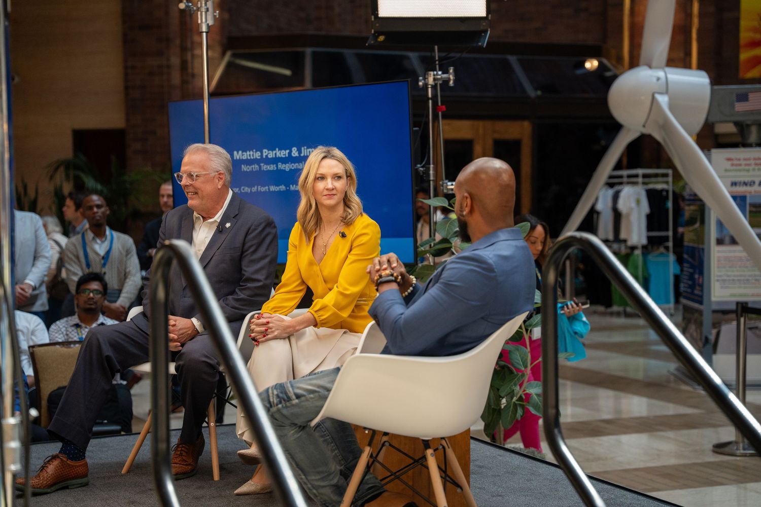 Three people sitting in chairs during a television interview setup with production equipment and blue backdrop.