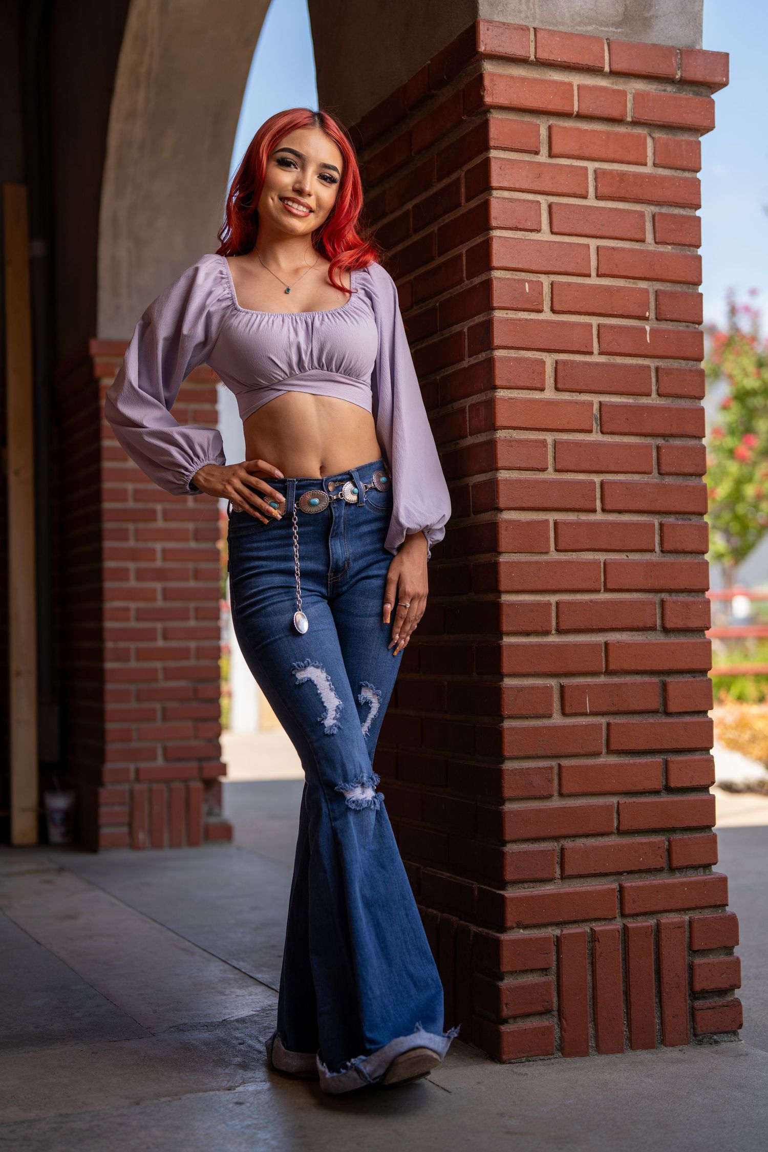 Someone with bright red hair poses against a brick pillar wearing a lavender crop top and flared distressed jeans.
