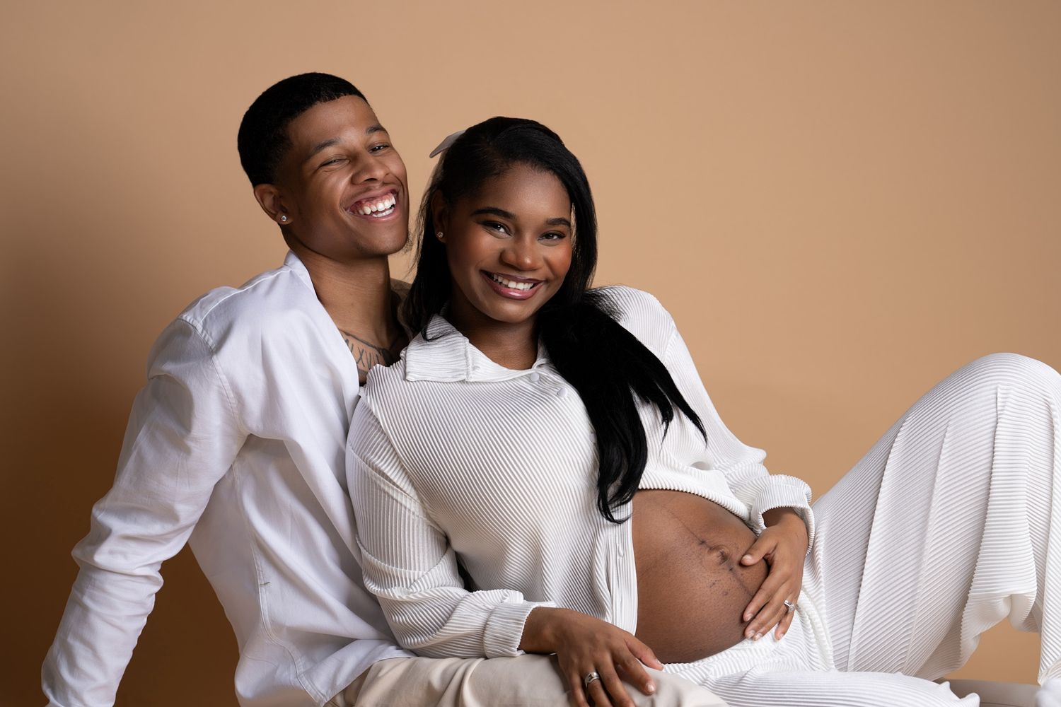 A couple in matching white outfits poses together for an intimate maternity photo against a tan backdrop.
