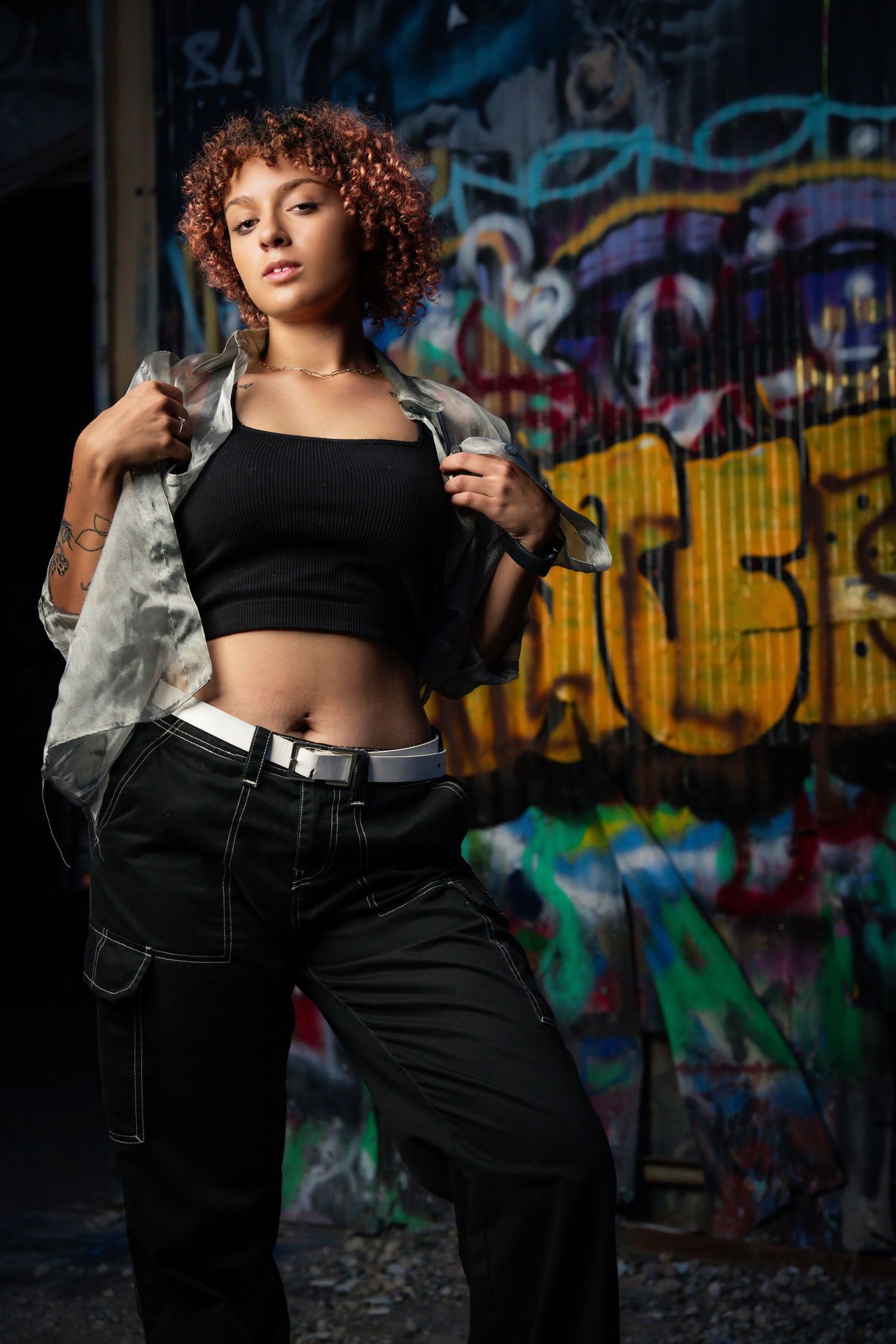 Fashion model in black crop top and baggy jeans poses against colorful graffiti wall in urban setting.
