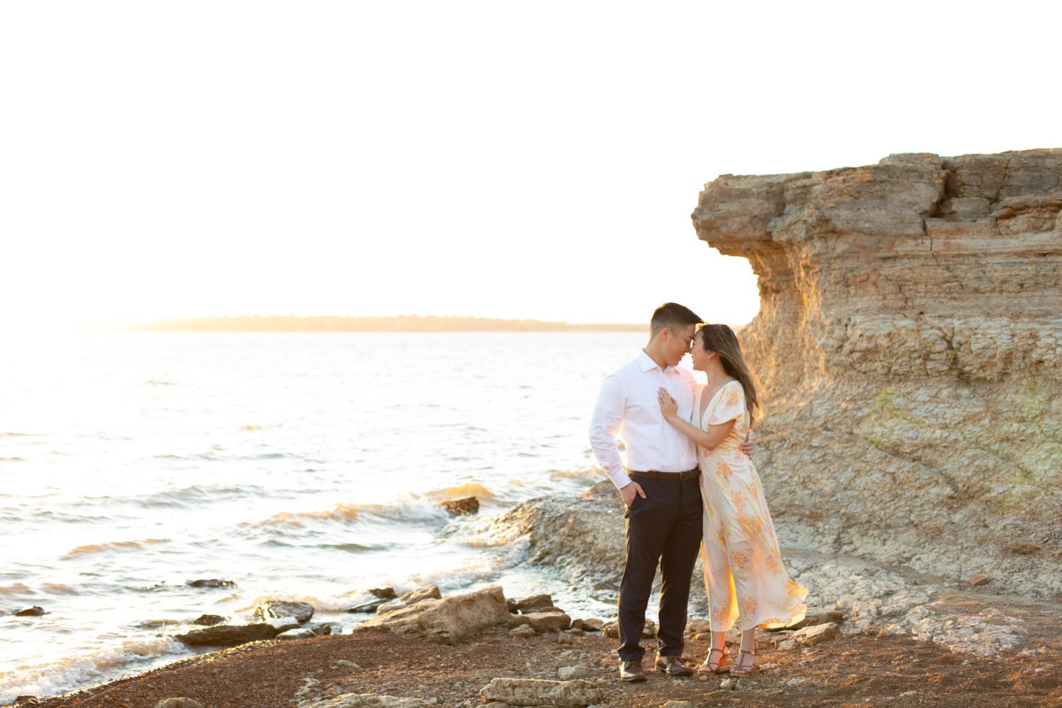 Romantic couple embraces on cliff edge overlooking ocean at sunset with rocky shoreline in the background.