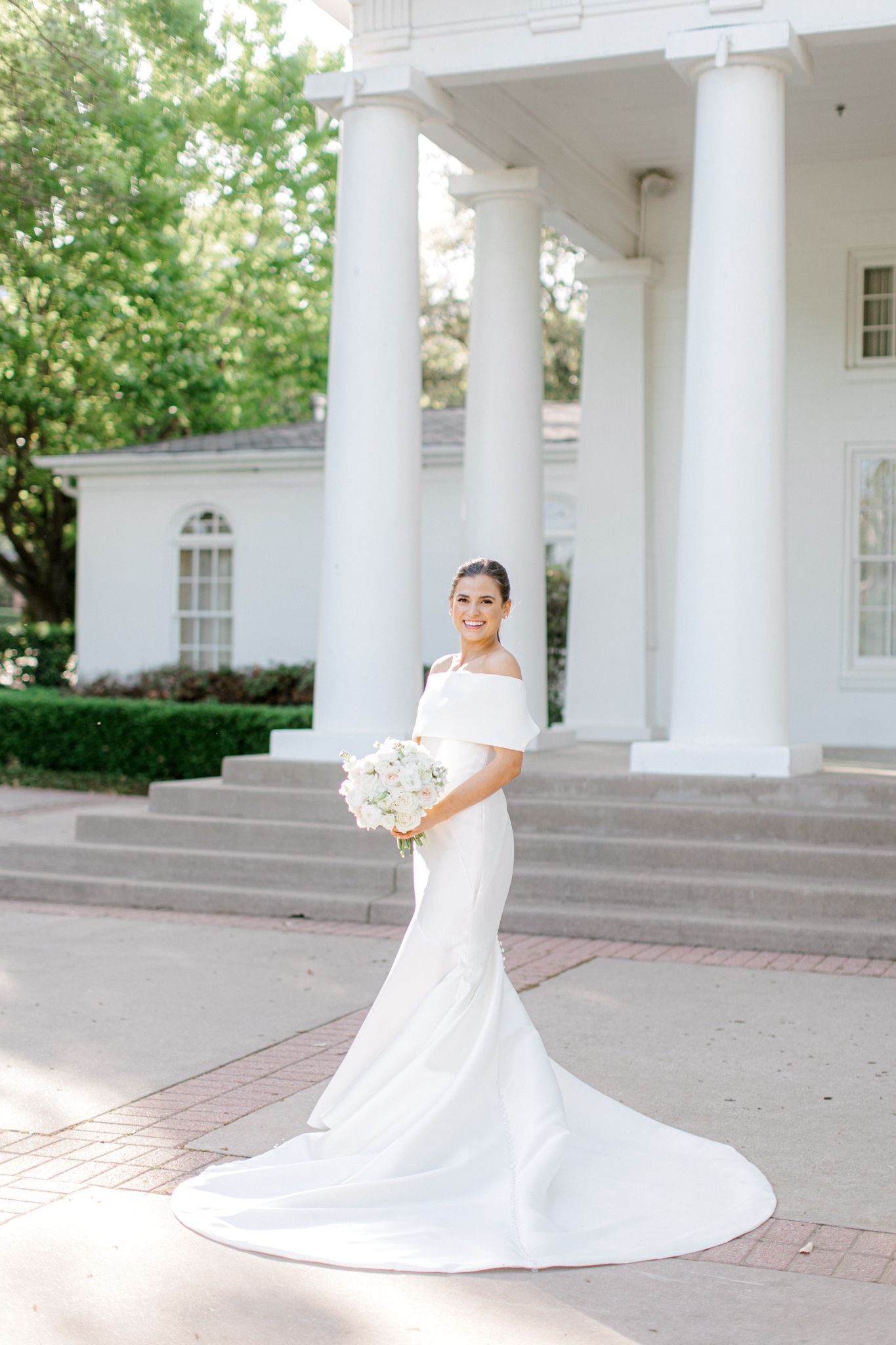 Elegant white wedding dress with cascading train billows on courtyard with classic white columns in the background.