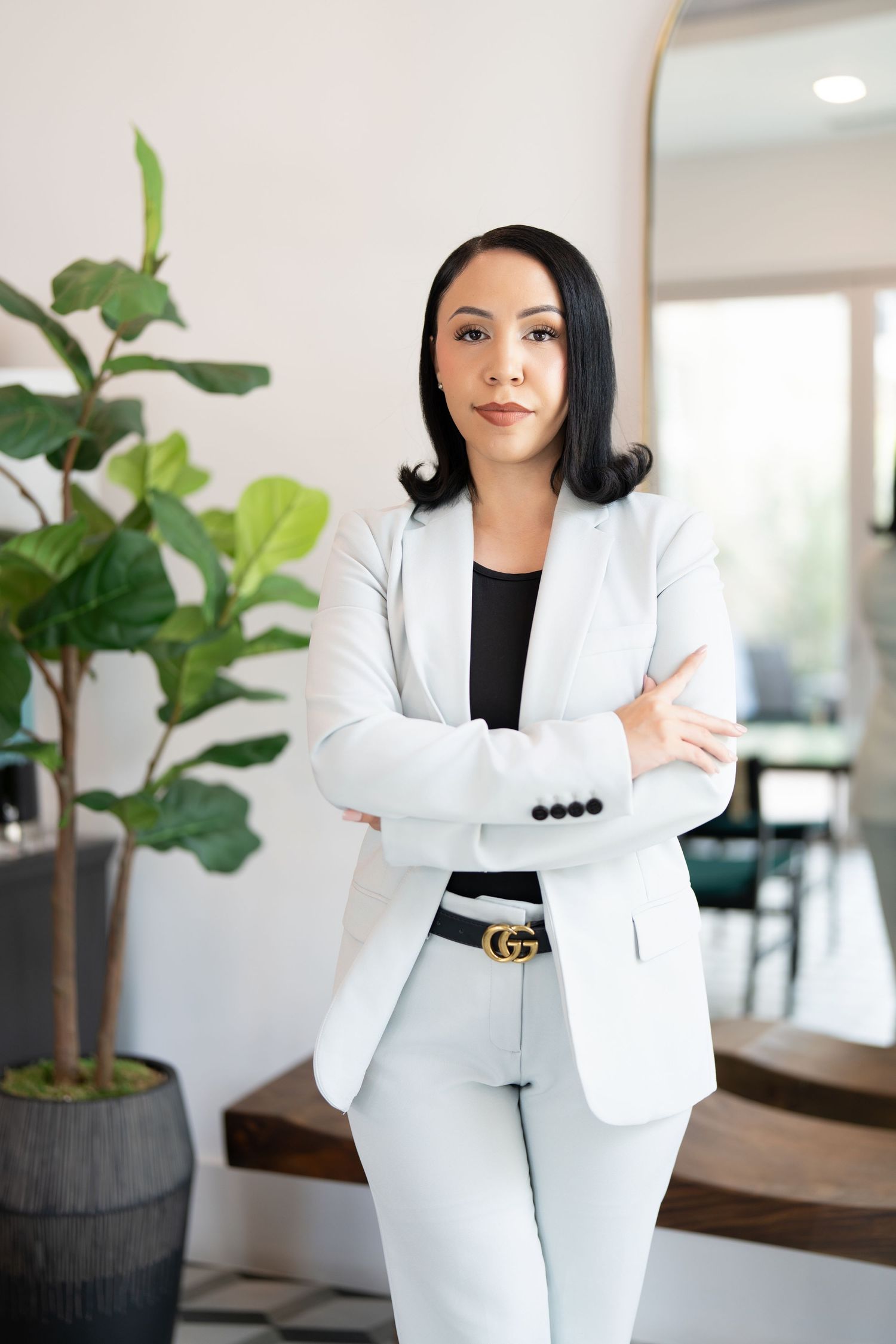 A stylish professional in a white pantsuit poses next to a decorative indoor plant.